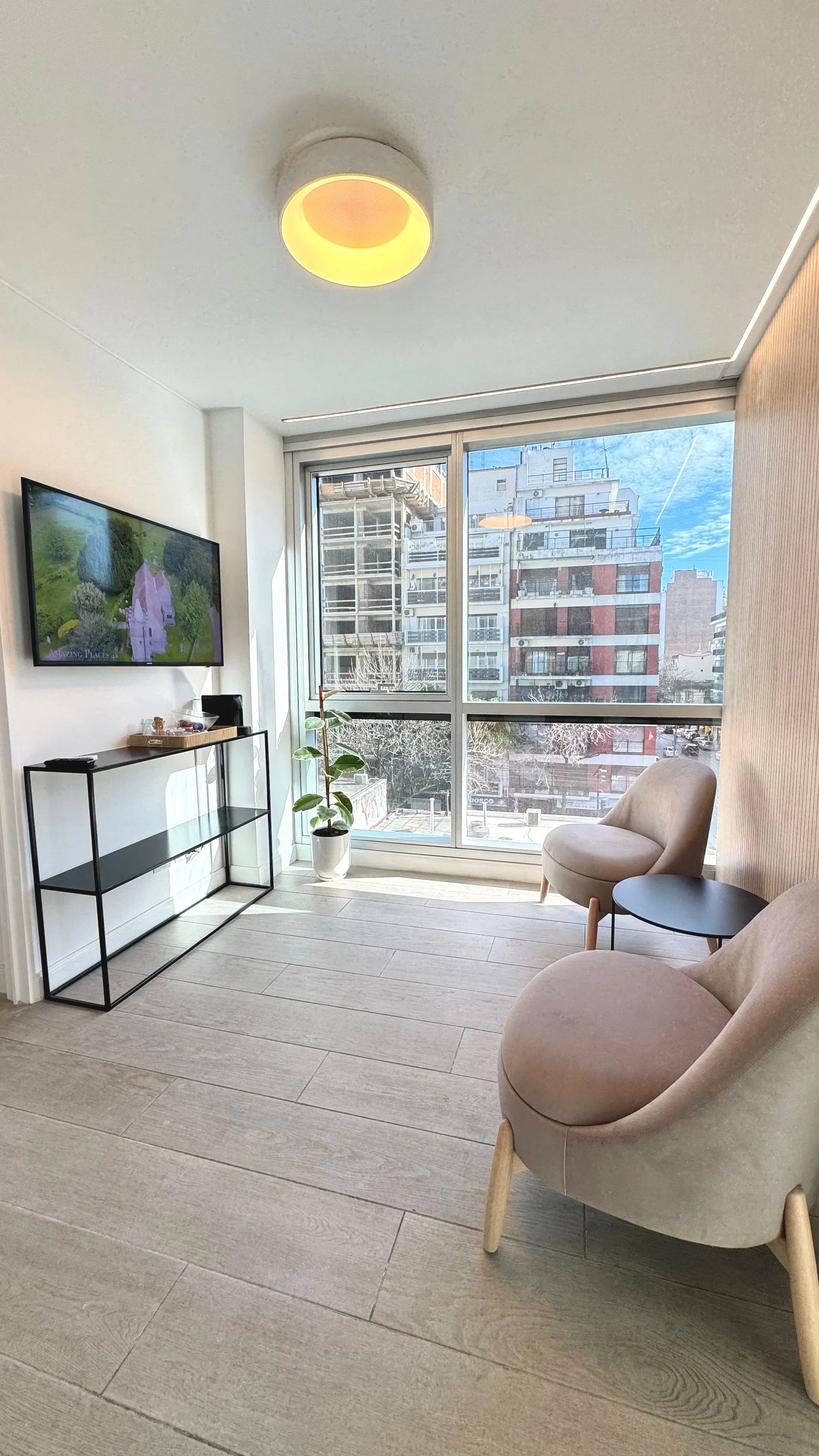Sala moderna con sillones beige, mesa redonda negra, televisión en la pared, planta en maceta, ventana grande con vista a edificios urbanos y cielo despejado.