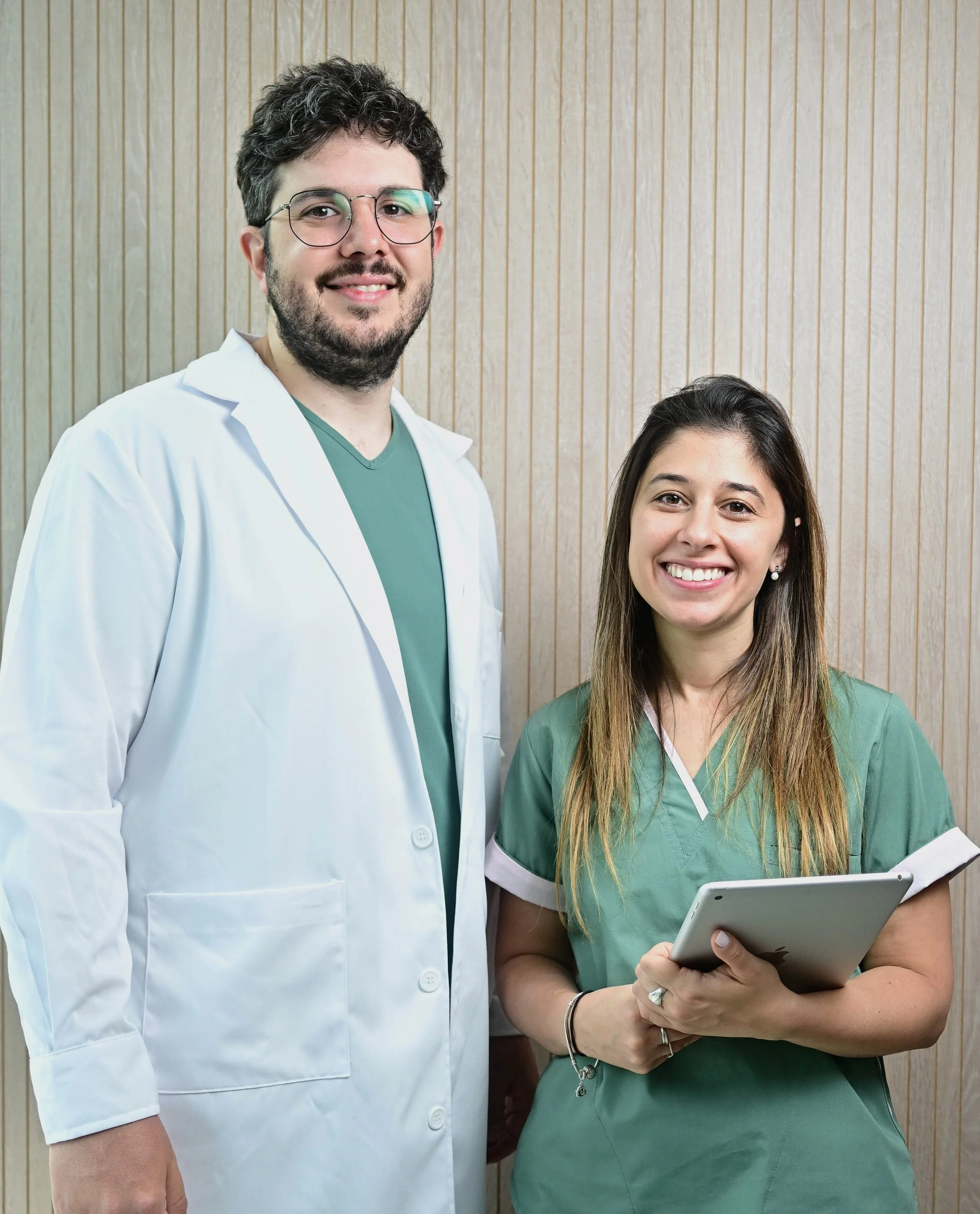 Dos profesionales de la salud, un hombre con bata blanca y una mujer con uniforme verde, posando en una clínica u hospital, sonriendo y mirando a la cámara.