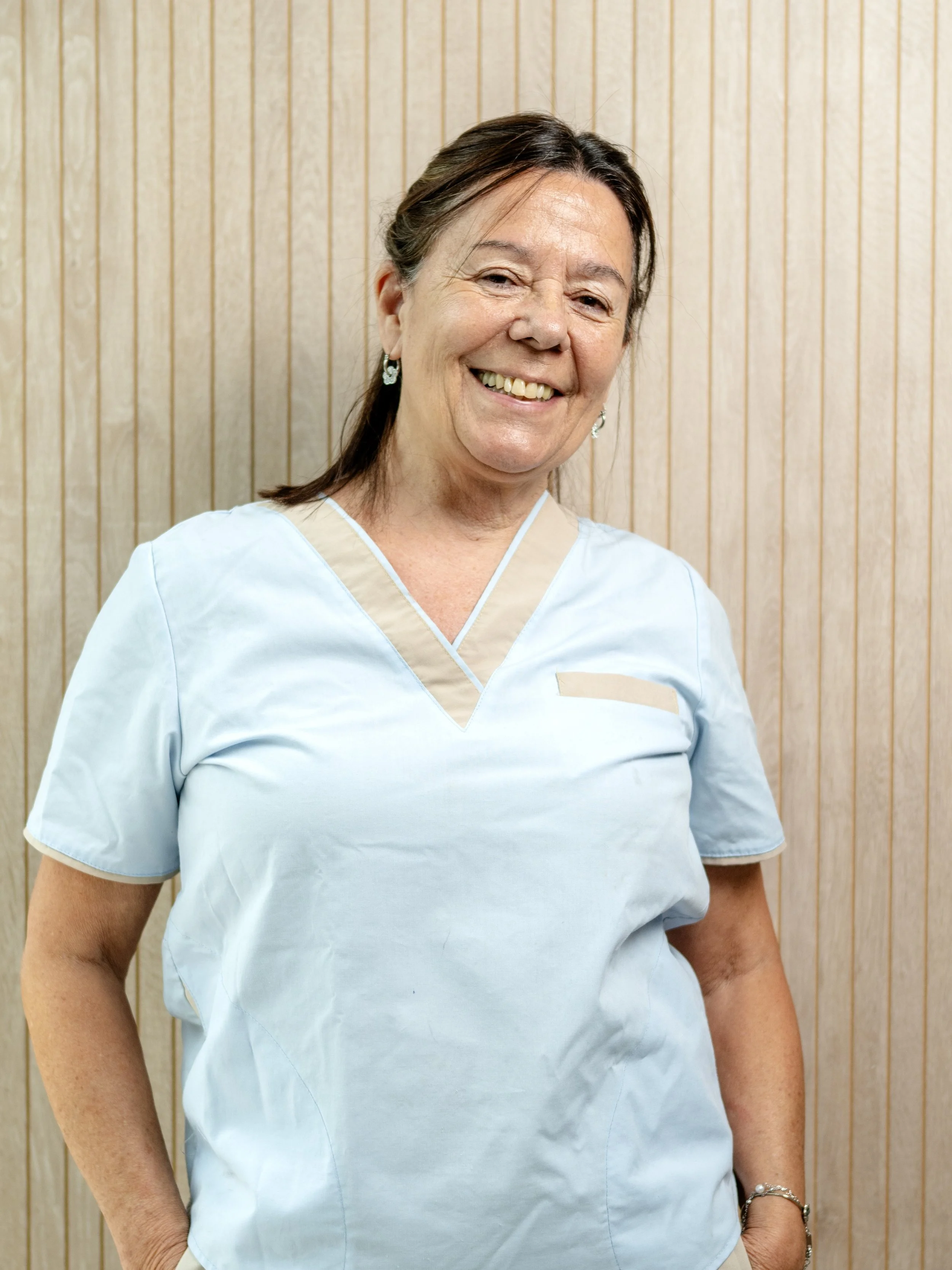 Una mujer de cabello castaño oscuro, con una sonrisa, usa uniforme de trabajo en tono celeste y fondo de madera vertical.