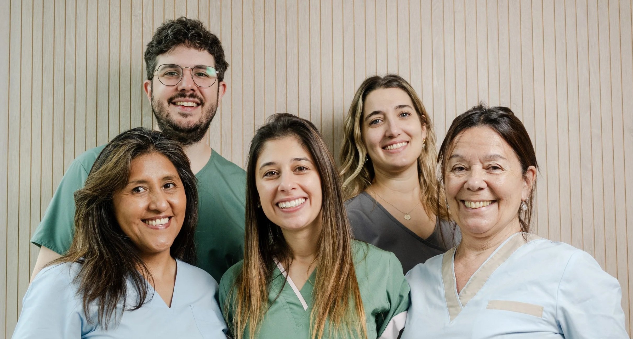 Grupo de seis profesionales de la salud sonriendo, usando batas y uniformes médicos, en un fondo de pared de madera.