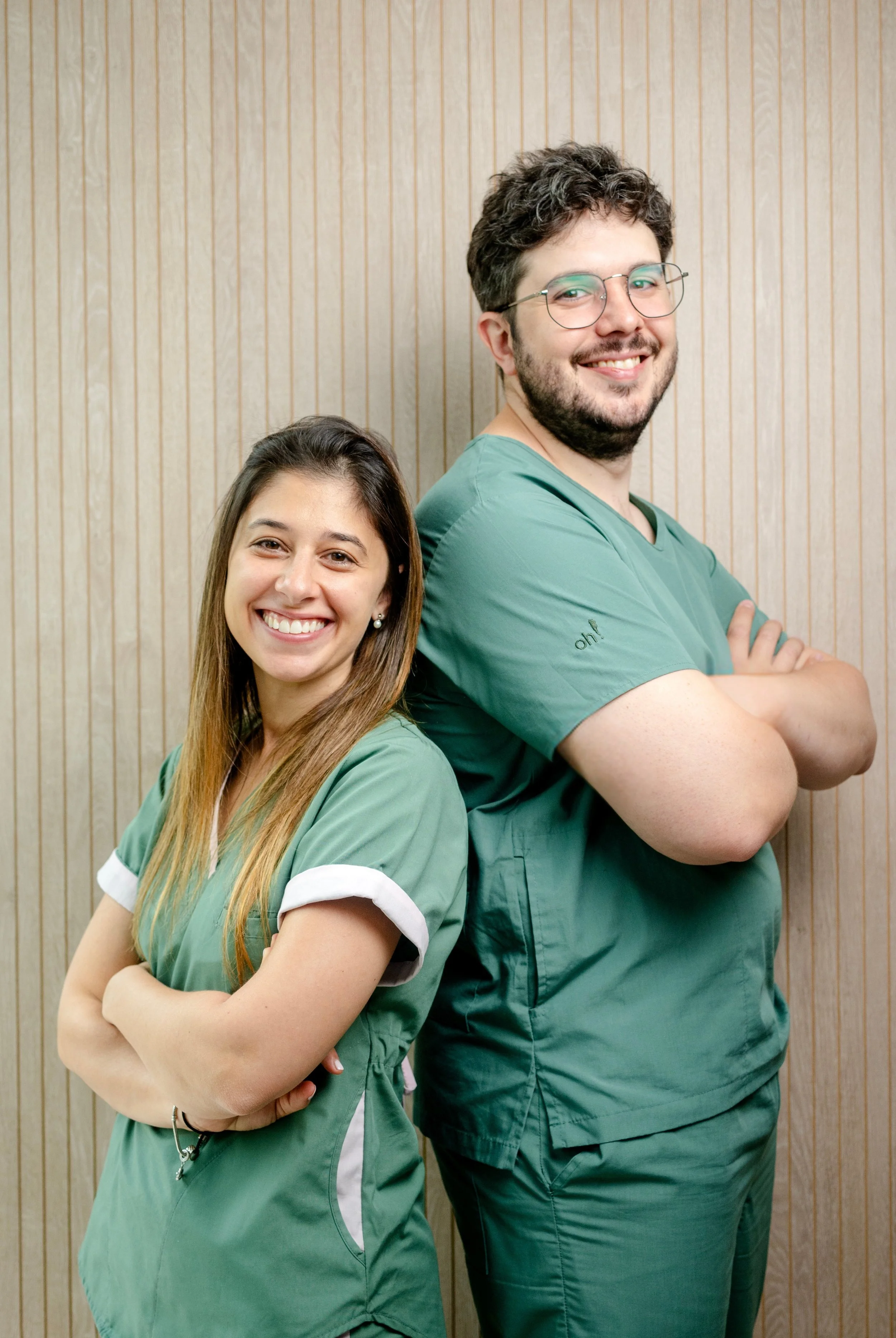 Un hombre y una mujer en uniformes de médico posando juntos contra una pared de madera, sonriendo y con los brazos cruzados.