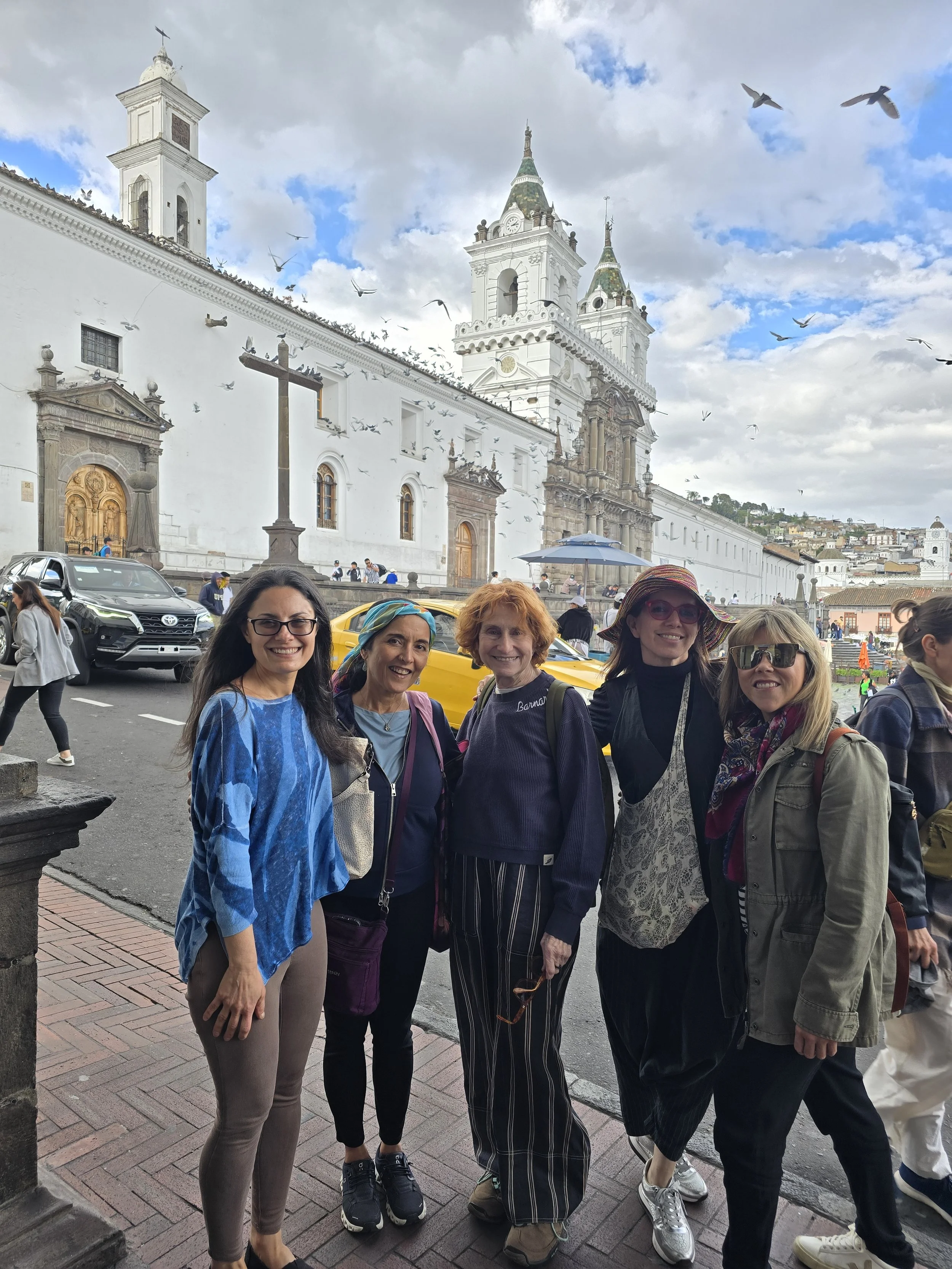 Group of five women in casual clothing posing on a city sidewalk in front of a historic white church with twin towers, flying pigeons, and a partly cloudy sky.