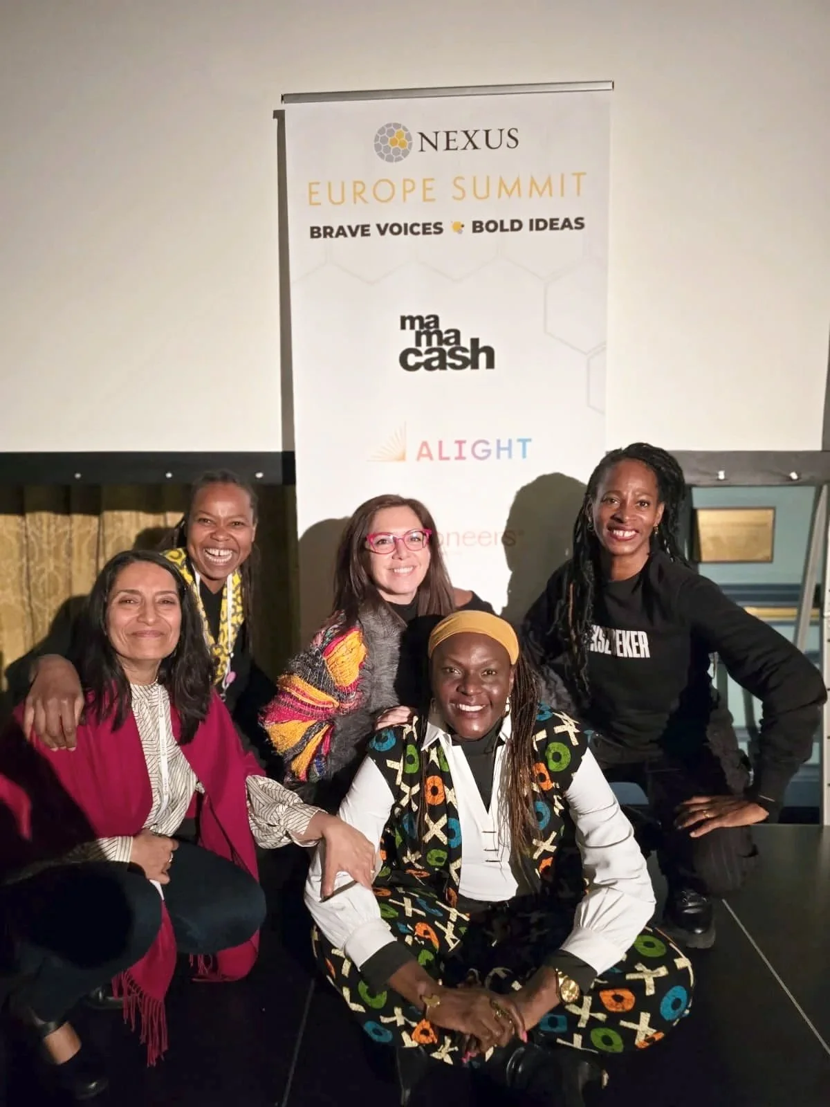 Group of five diverse women smiling and posing together at the Nexus Europe Summit event, standing and sitting in front of a large banner.