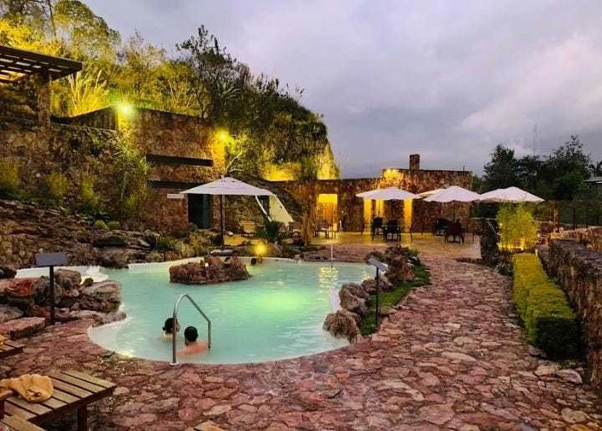 People swimming in a hot spring pool surrounded by rocks, with terrace seating, umbrellas, and lush greenery at dusk.