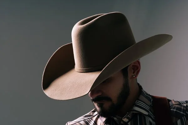 A person wearing a large cowboy hat and a striped shirt, with a beard, looking down.