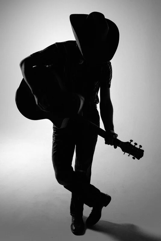 Silhouette of a person wearing a cowboy hat, carrying a guitar on their back, and holding another guitar in their hand, against a plain background.