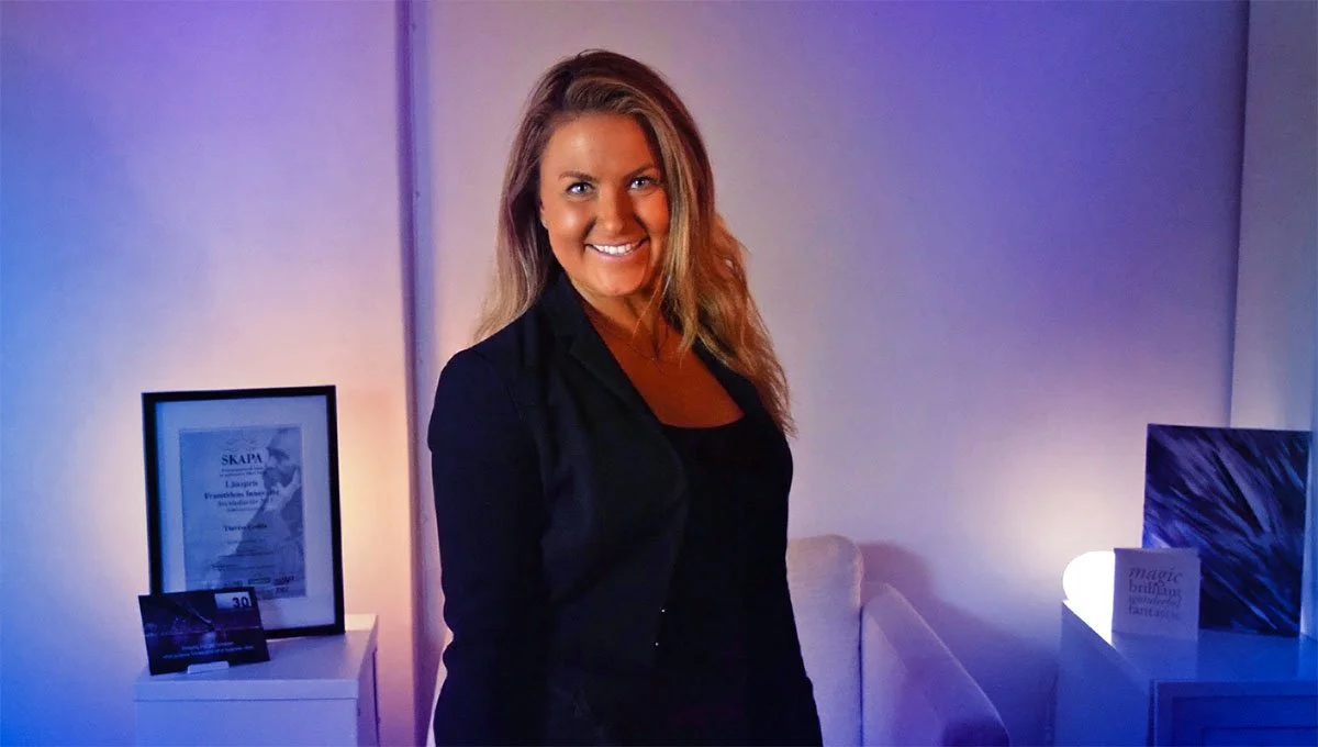 A smiling woman with long wavy hair wearing a black blazer standing in a room with colorful lighting and artwork.