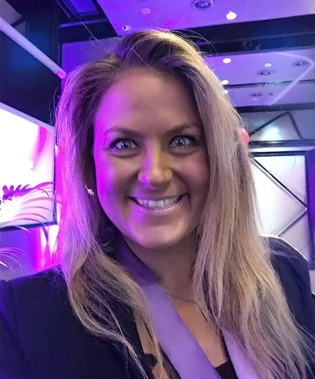 A woman with long blonde hair and blue eyes smiling at the camera in a setting with purple and blue lighting, possibly at an event or conference.