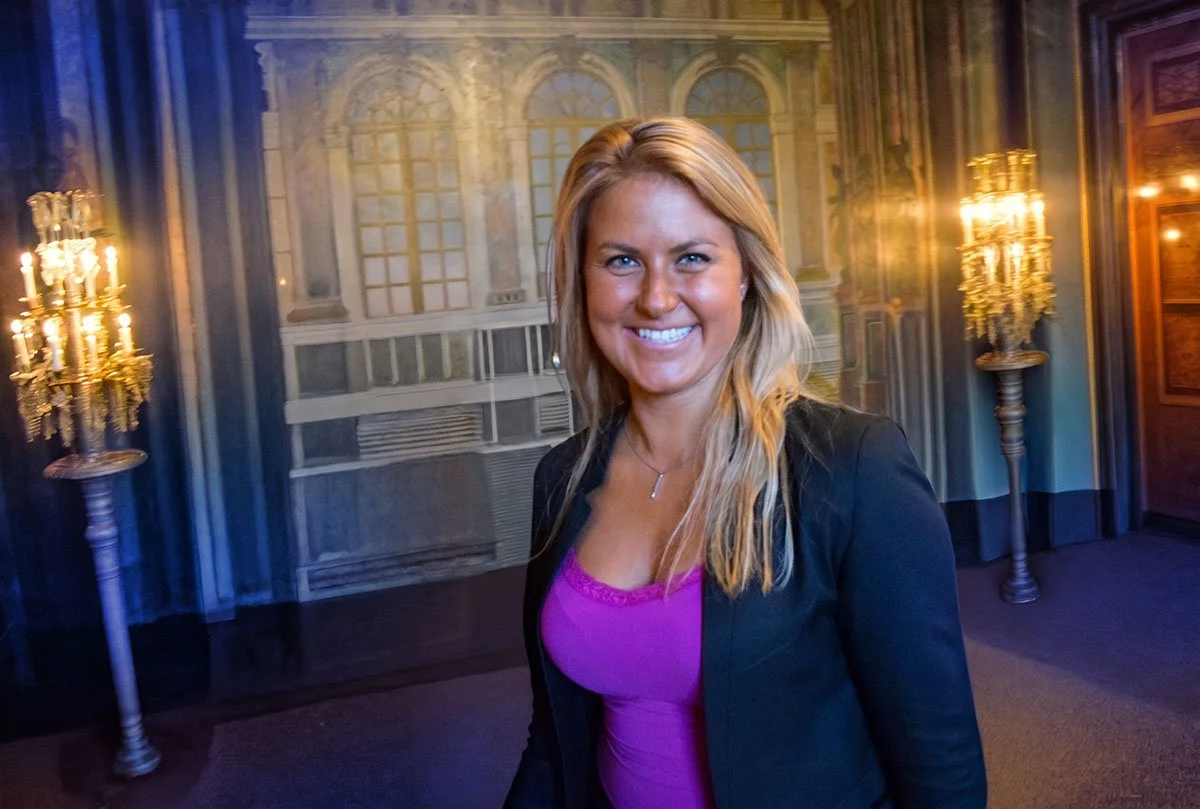 A smiling woman with blonde hair wearing a purple top and black blazer standing in a room with ornate décor, including chandeliers and decorative wall panels.