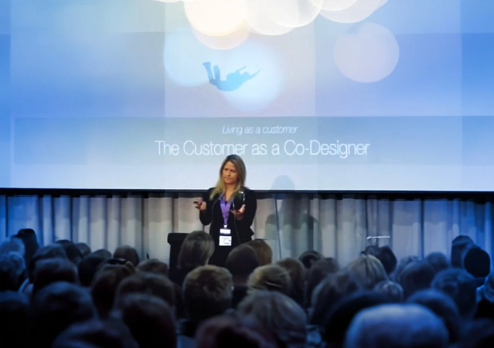 A woman giving a presentation at a conference, with a large screen behind her displaying the title "Living as a customer: The Customer as a Co-Designer." The audience is seated in front of her, and the room is dimly lit.