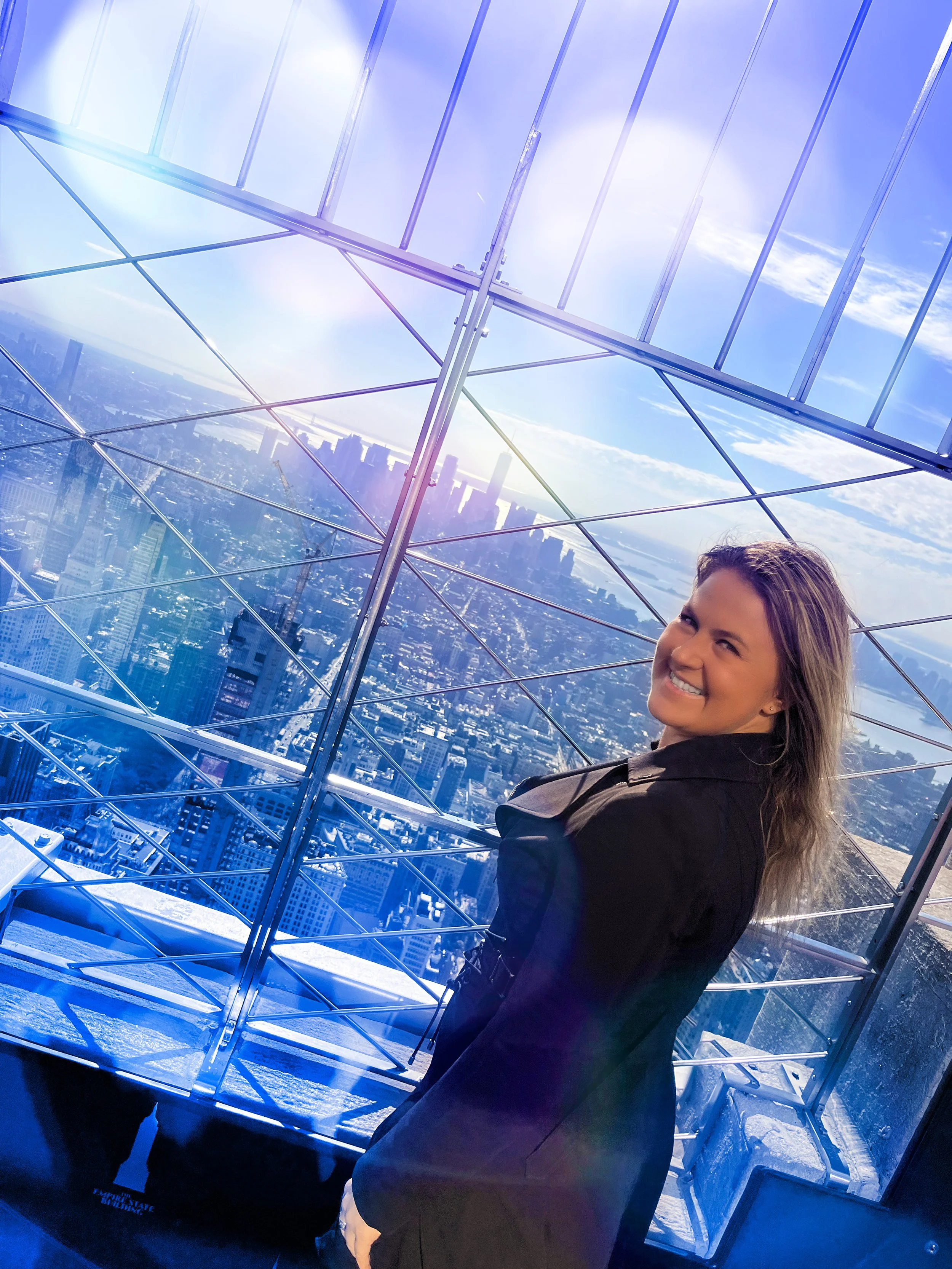 A smiling woman with long hair, dressed in a black jacket, standing inside a glass observation deck with a city skyline and blue sky visible outside.