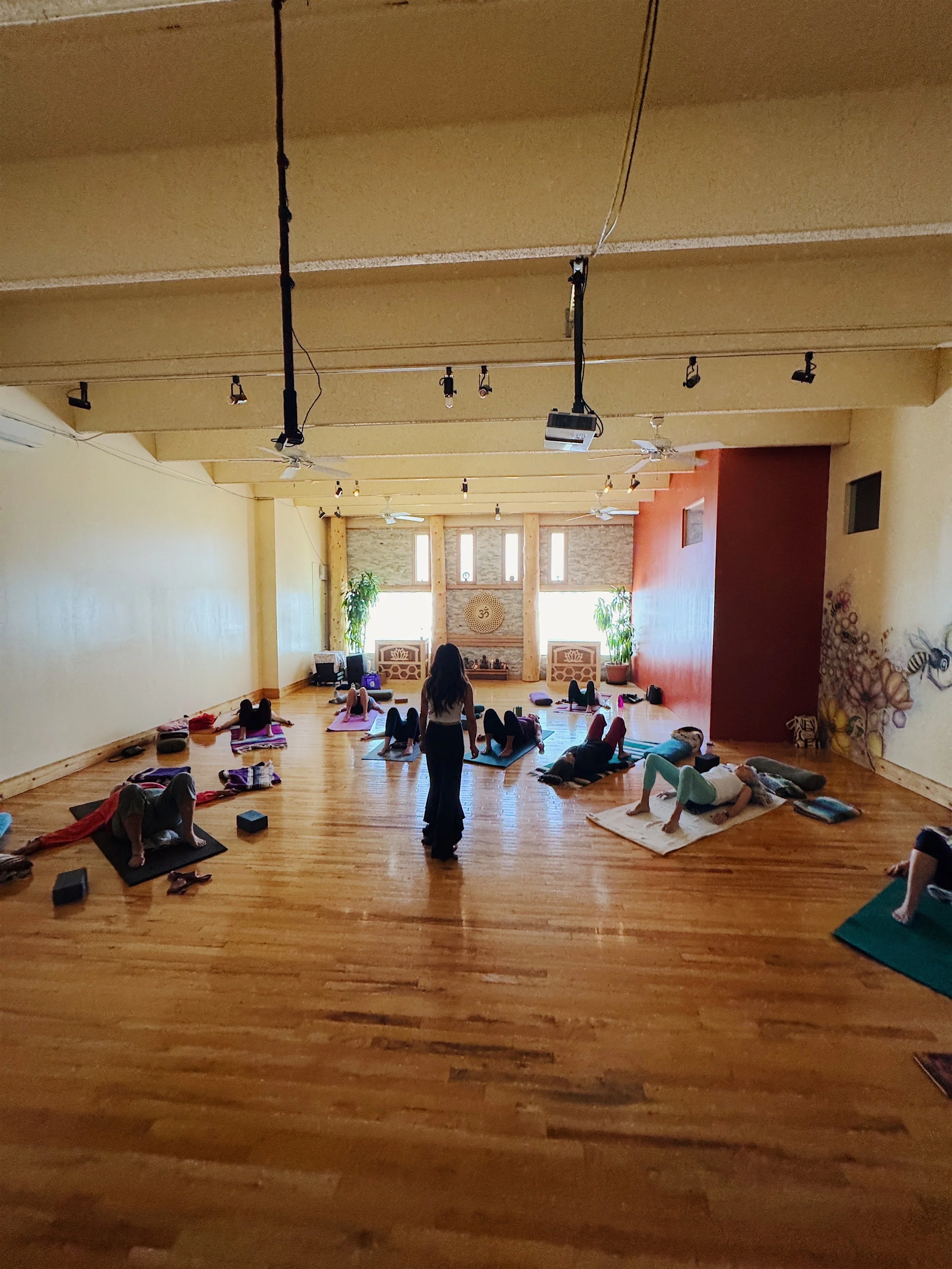 A group yoga class taking place in a spacious wooden-floored room with large windows, yoga mats, and participants practicing stretching poses, with an instructor observing.