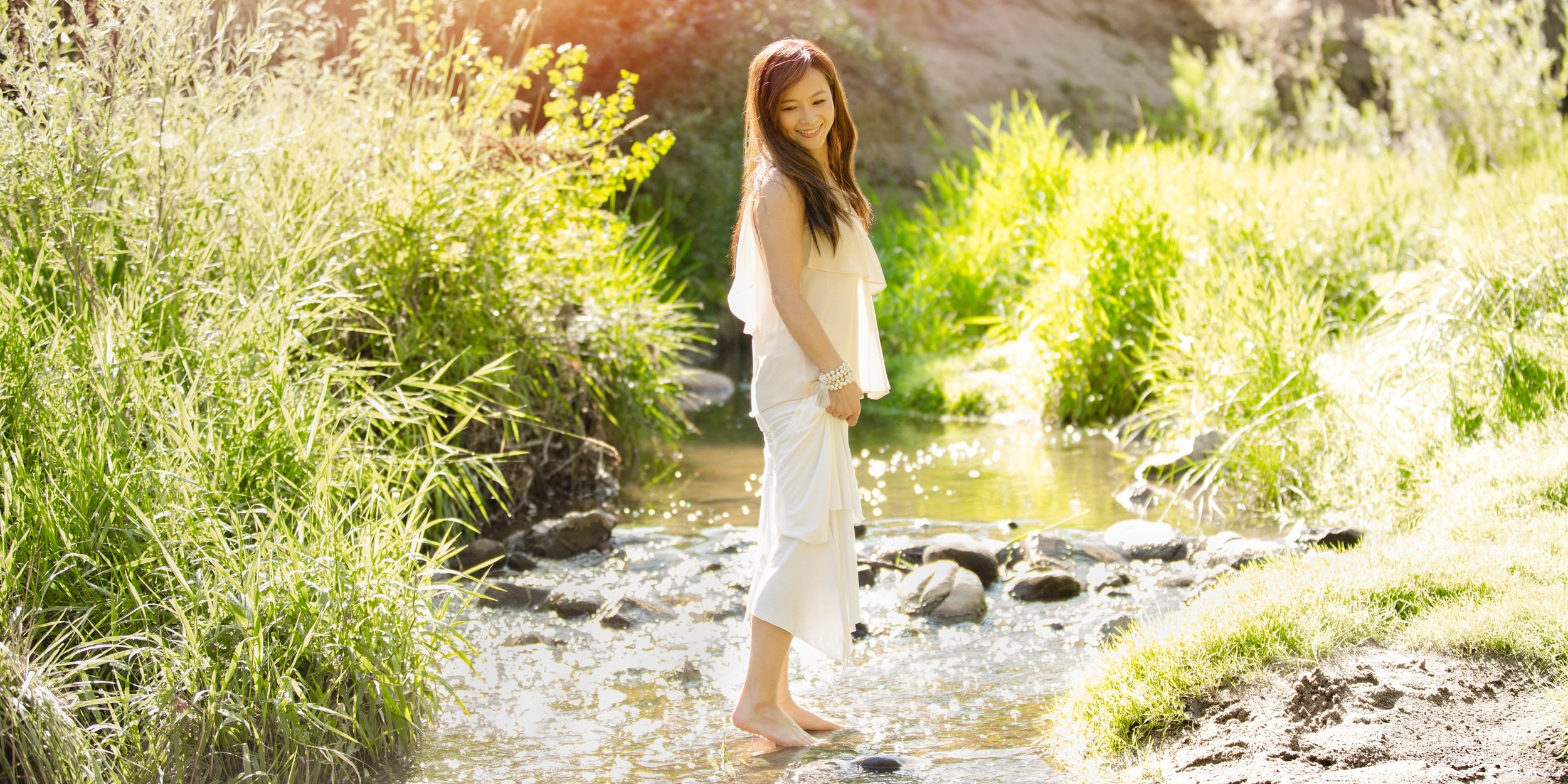 A woman in a white dress standing in a shallow stream surrounded by lush green grass and plants, smiling at the camera on a sunny day.