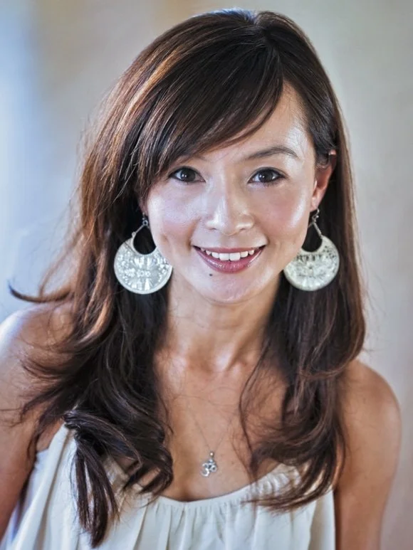 A woman smiling at the camera with long brown hair, wearing large silver earrings and a cream-colored top.