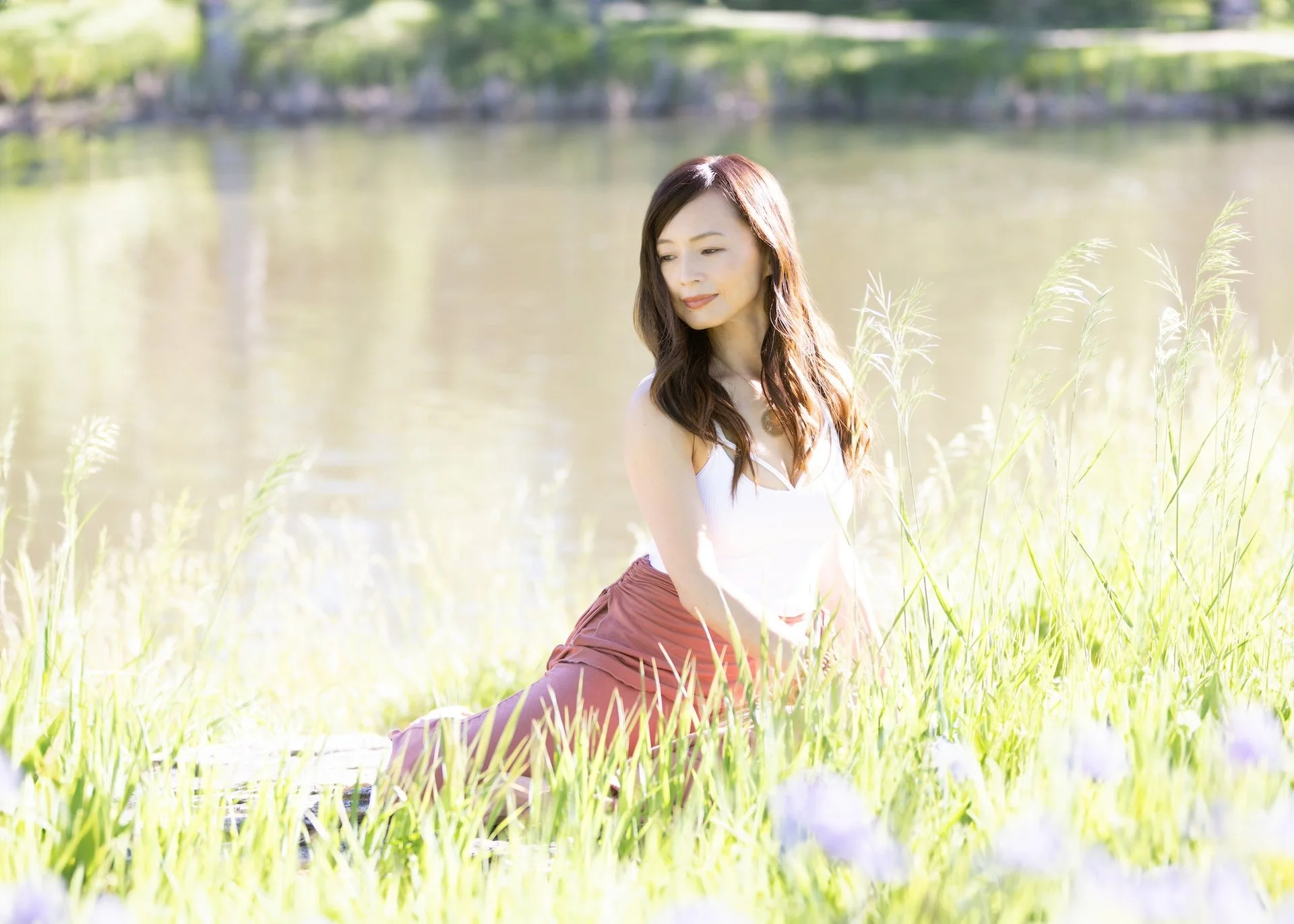 A woman kneels in a grassy field near a body of water, wearing a white top and reddish pants, with her eyes closed and a serene expression.
