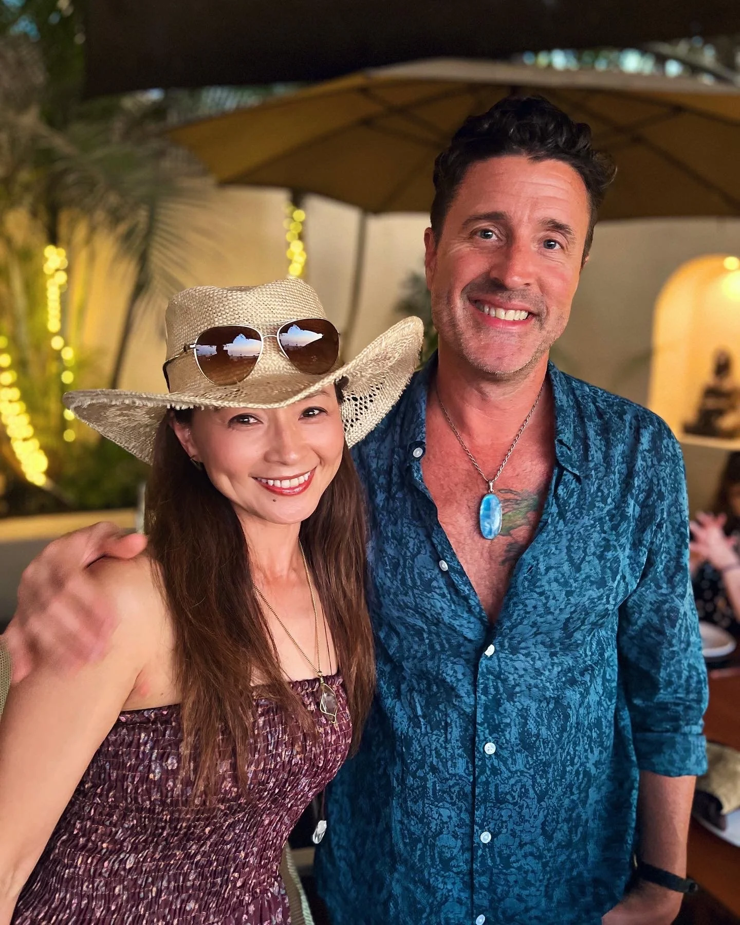 A smiling woman wearing a straw hat with sunglasses on it, and a man with dark hair and a blue patterned shirt, standing together at an outdoor gathering, with warm lighting and string lights in the background.