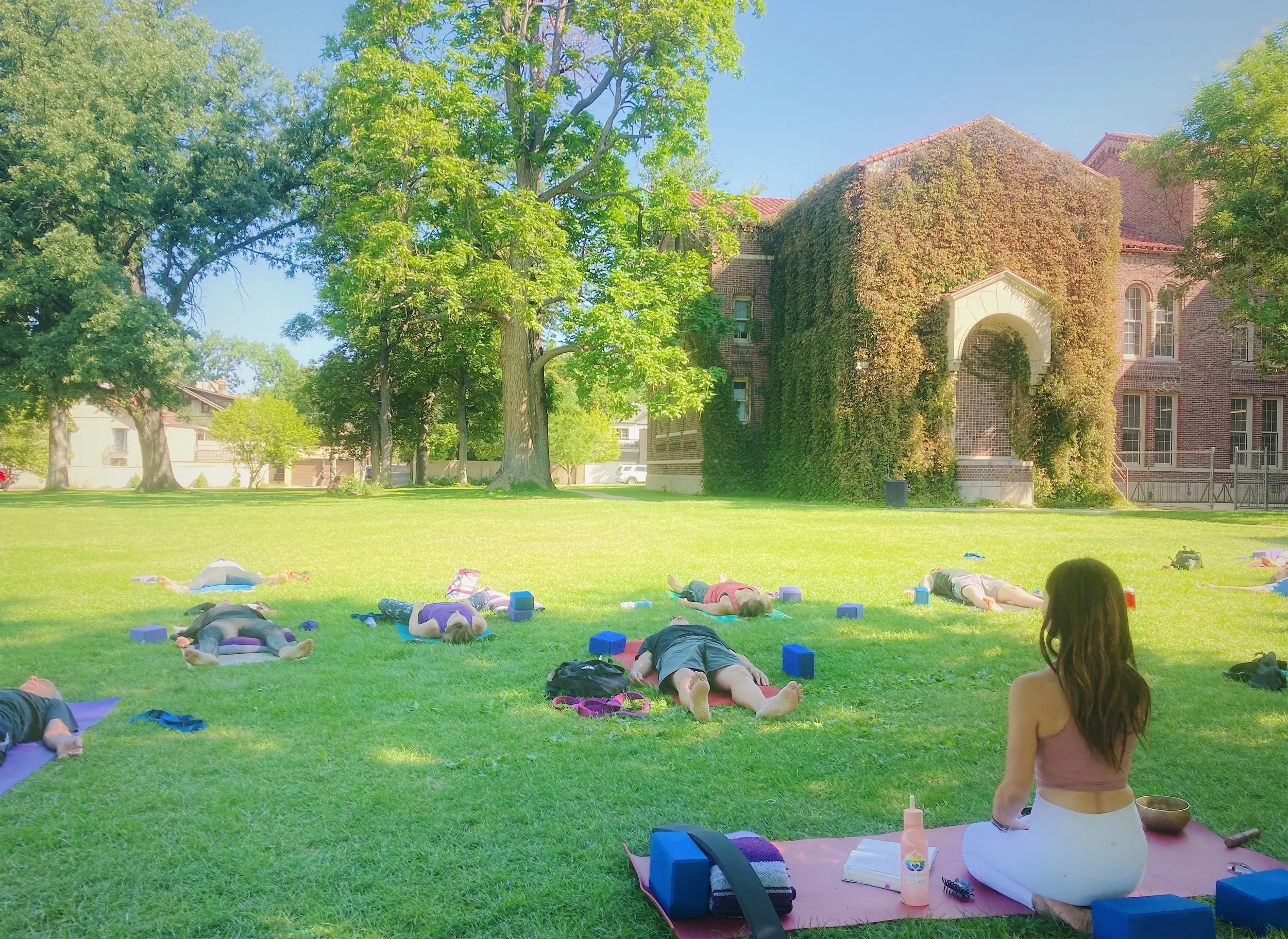 People participating in an outdoor yoga class on a grassy area with trees and a brick building covered in ivy in the background. A woman with long dark hair, wearing a pink top and white pants, is sitting on a yoga mat facing away from the camera, with various yoga accessories around her.