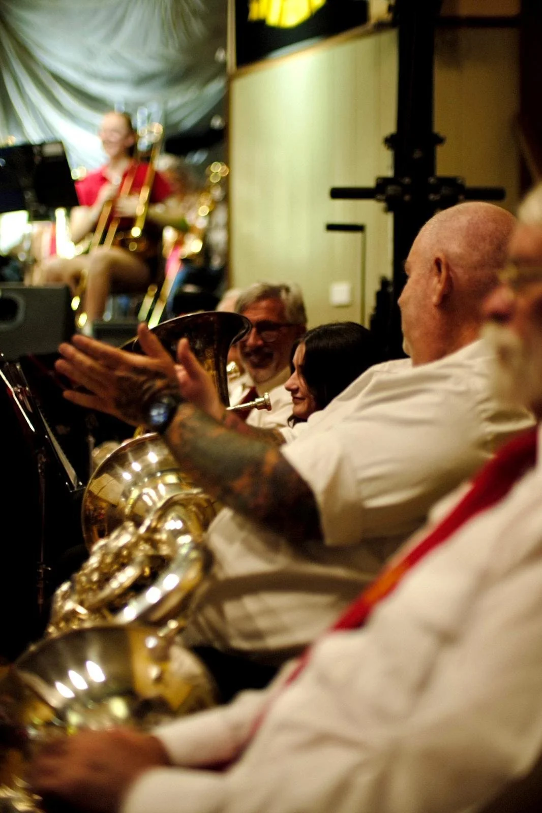 A group of musicians, including a person with tattoos on their arm, sitting together with their brass instruments, watching a performer on stage in a lively indoor setting.