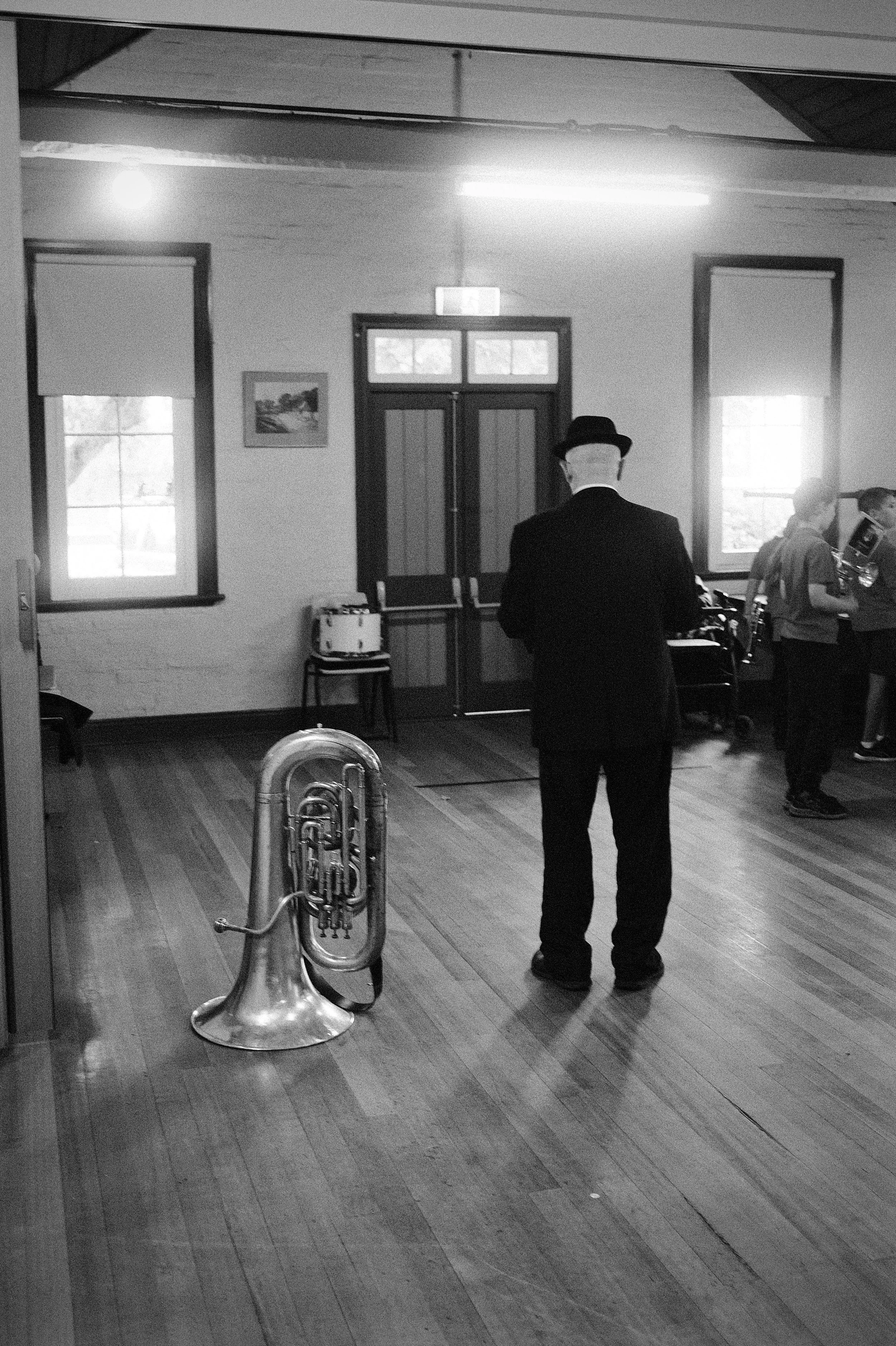 A man in a suit and bowler hat standing in a room with wooden floors, with a sousaphone instrument on the floor nearby. There are two windows on the wall.