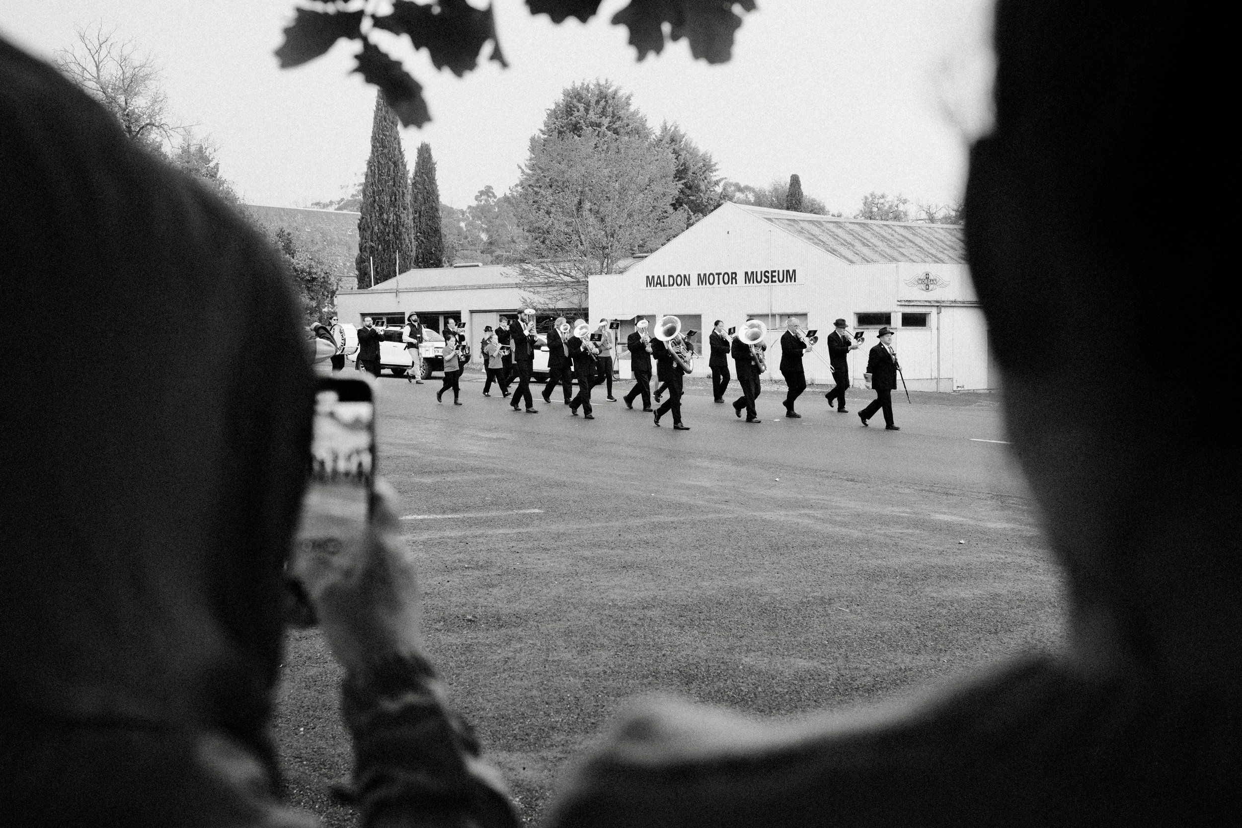 A marching band performing in front of the Maldon Motor Museum, viewed through the frame of two spectators.