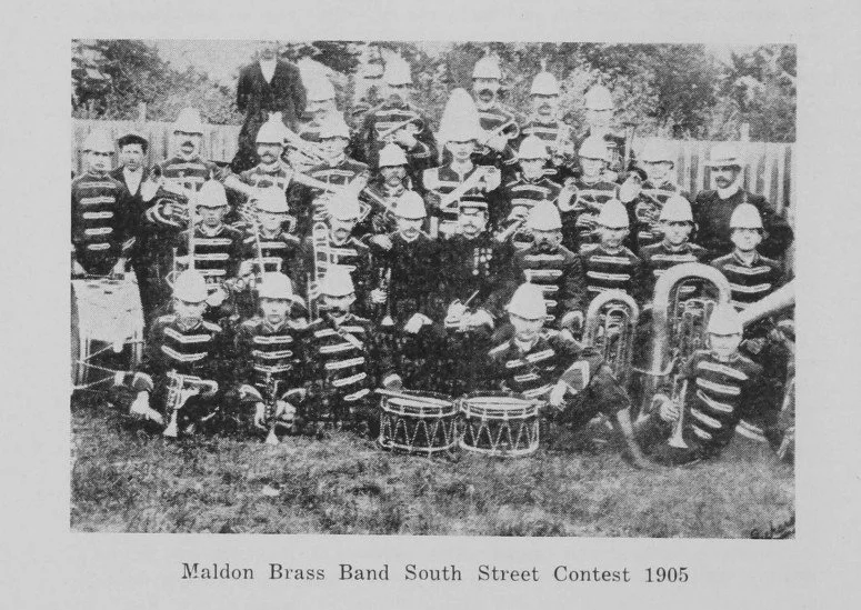 Black and white photograph of the Maldon Brass Band at the South Street Contest in 1905, showing a group of children and adults with band instruments, outdoors with a fence and trees in the background.