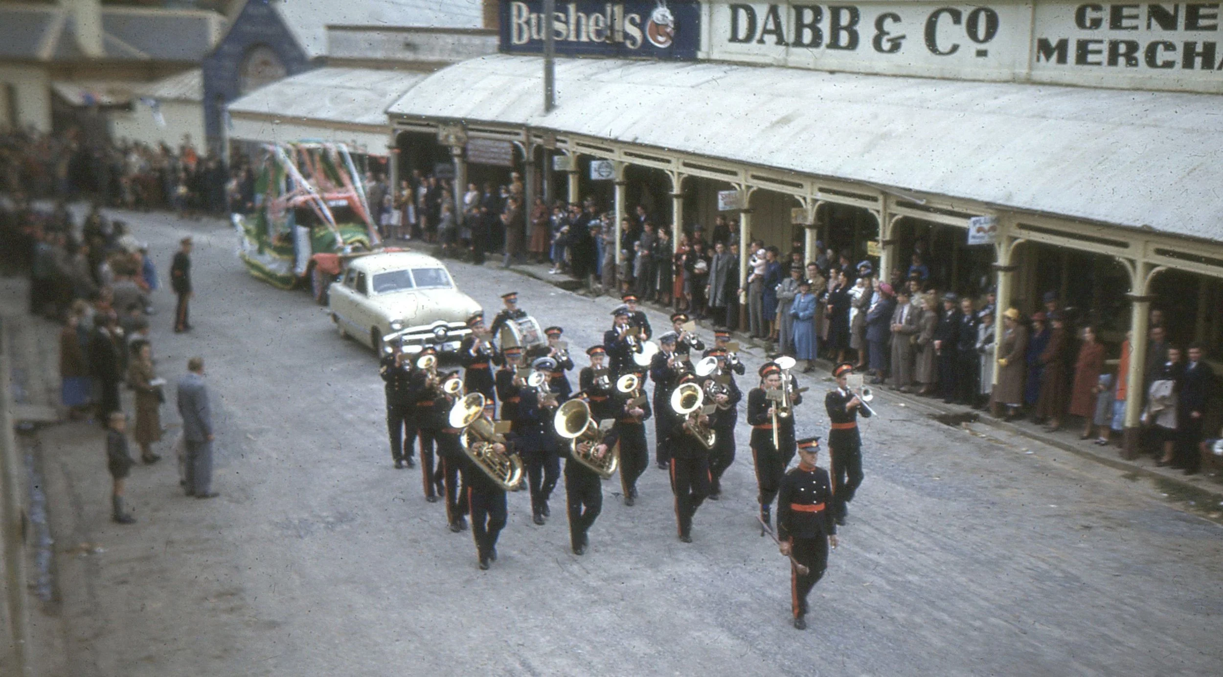 A marching band in uniform parades down a city street lined with onlookers, vintage cars, and storefronts from an earlier era.