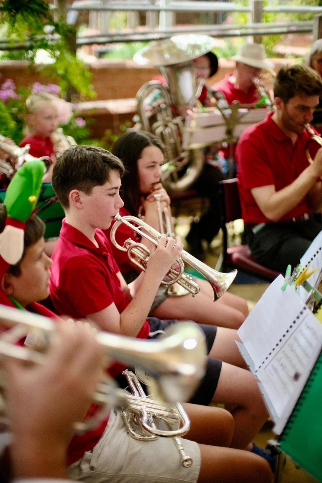 Children and adults playing brass and wind instruments in a band, seated indoors, some in festive holiday attire.