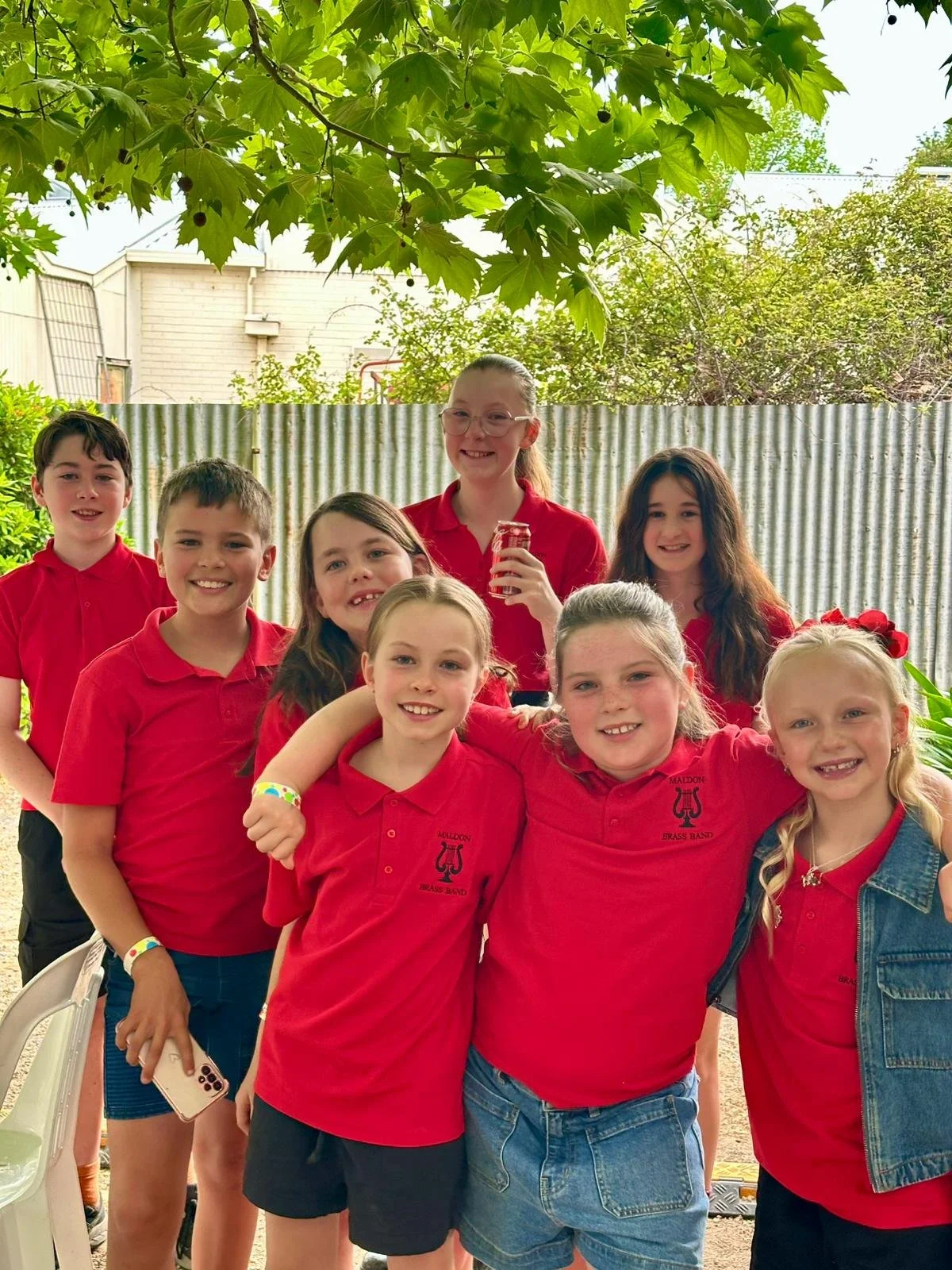 A group of children in red shirts posing outdoors, some with arms around each other, smiling at the camera.