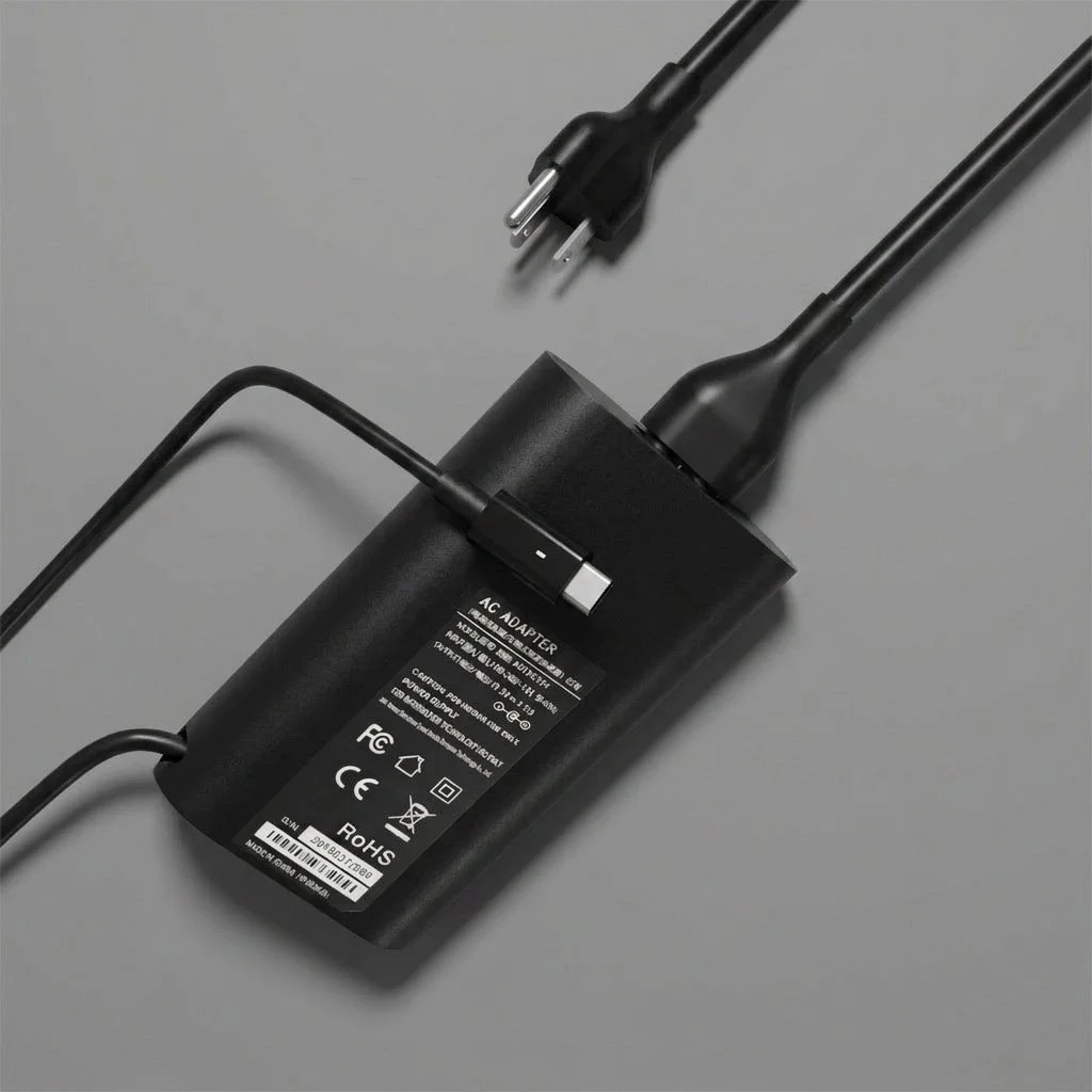 A black AC power adapter with a two-prong plug and a cord, placed on a gray surface.