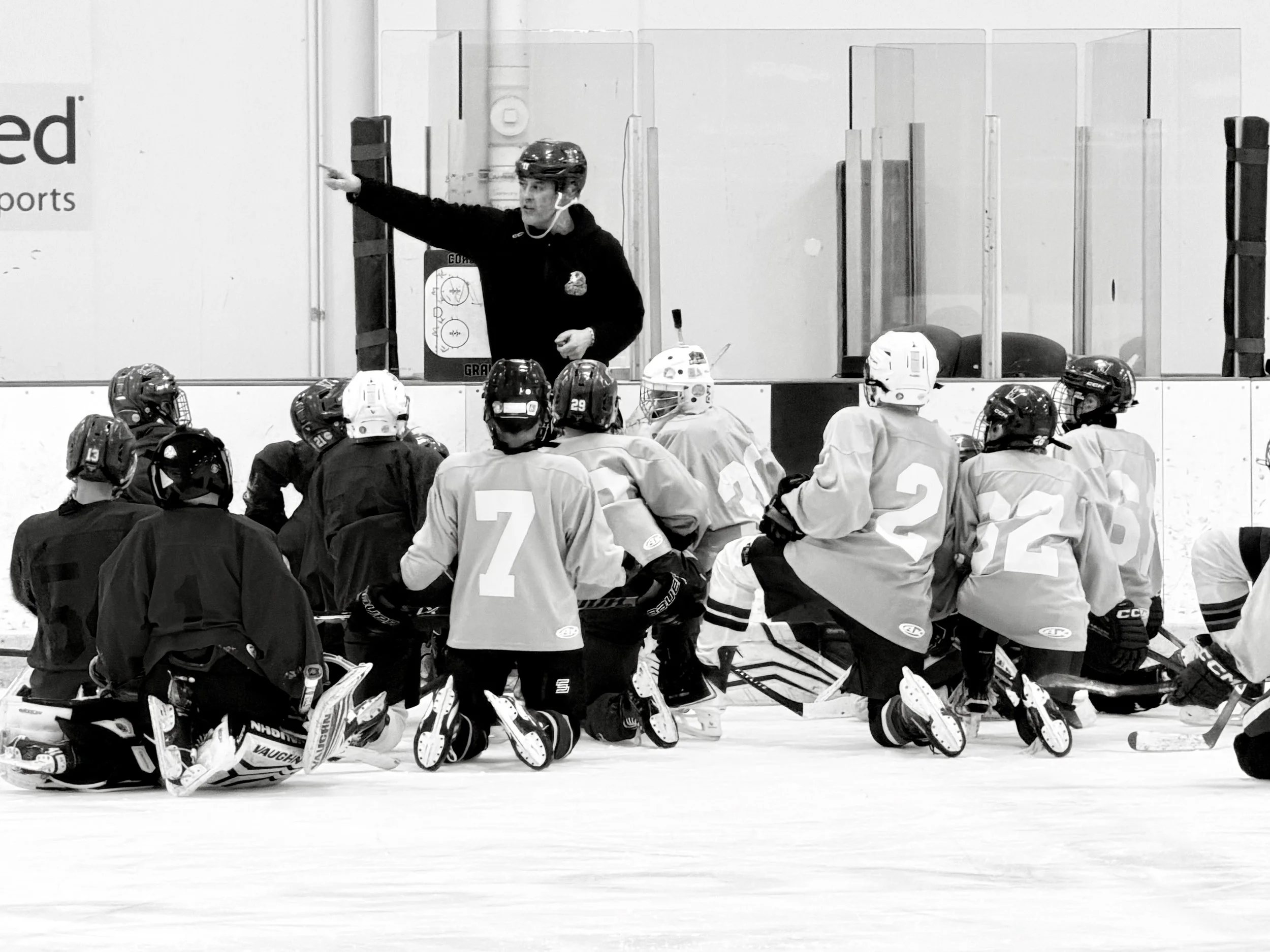 A hockey coach is giving instructions during a practice or game, with players kneeling on the ice listening attentively.