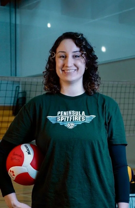 A young woman with curly dark hair smiling, holding a red and white volleyball with 'Mikasa' and a maple leaf logo, in a gymnasium with a volleyball net in the background, wearing a green t-shirt that says 'PENINSULA SPITFIRES'.
