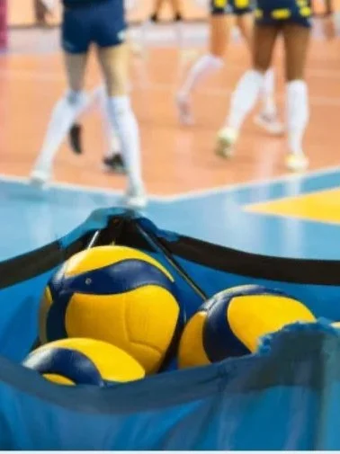 A blue sports bag filled with yellow and blue volleyballs on an indoor court, with volleyball players in uniform in the background.