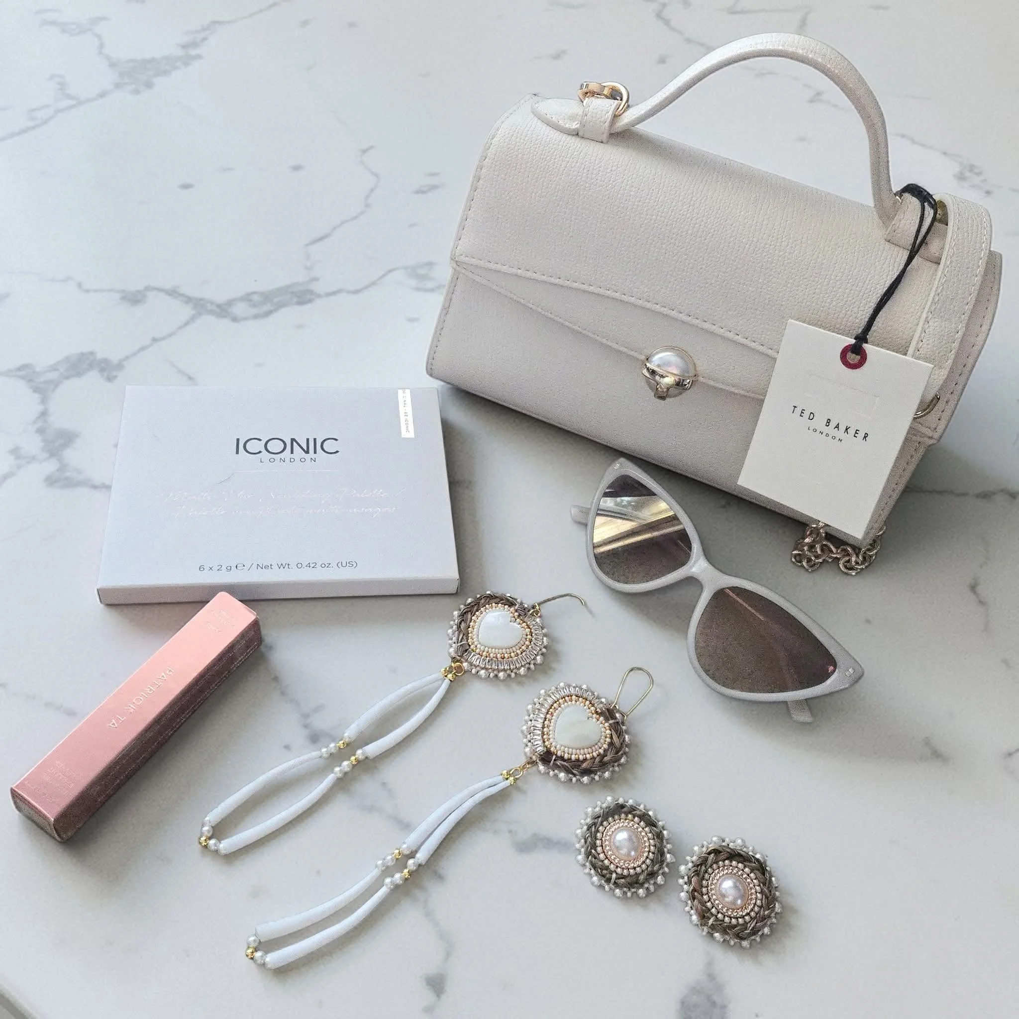 A white Ted Baker handbag, a pair of white sunglasses, luxury beaded earrings with white and pearl accents, a Marie Antoinette pink lipstick, a box of ICONIC London makeup blush, on a white marble surface.