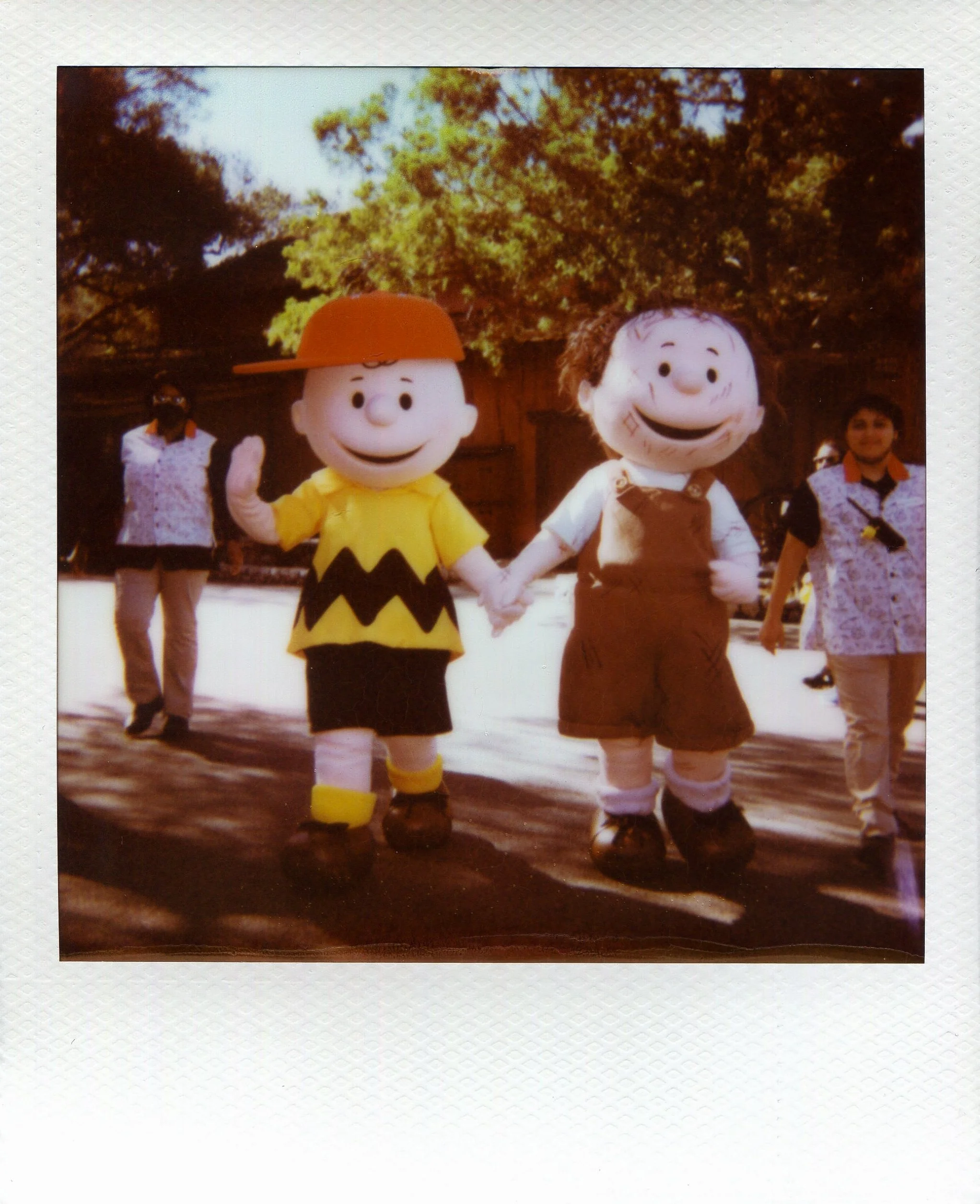 Costumed characters of Charlie Brown and Lucy holding hands, walking outdoors on a sunny day, with two people dressed casually in the background.