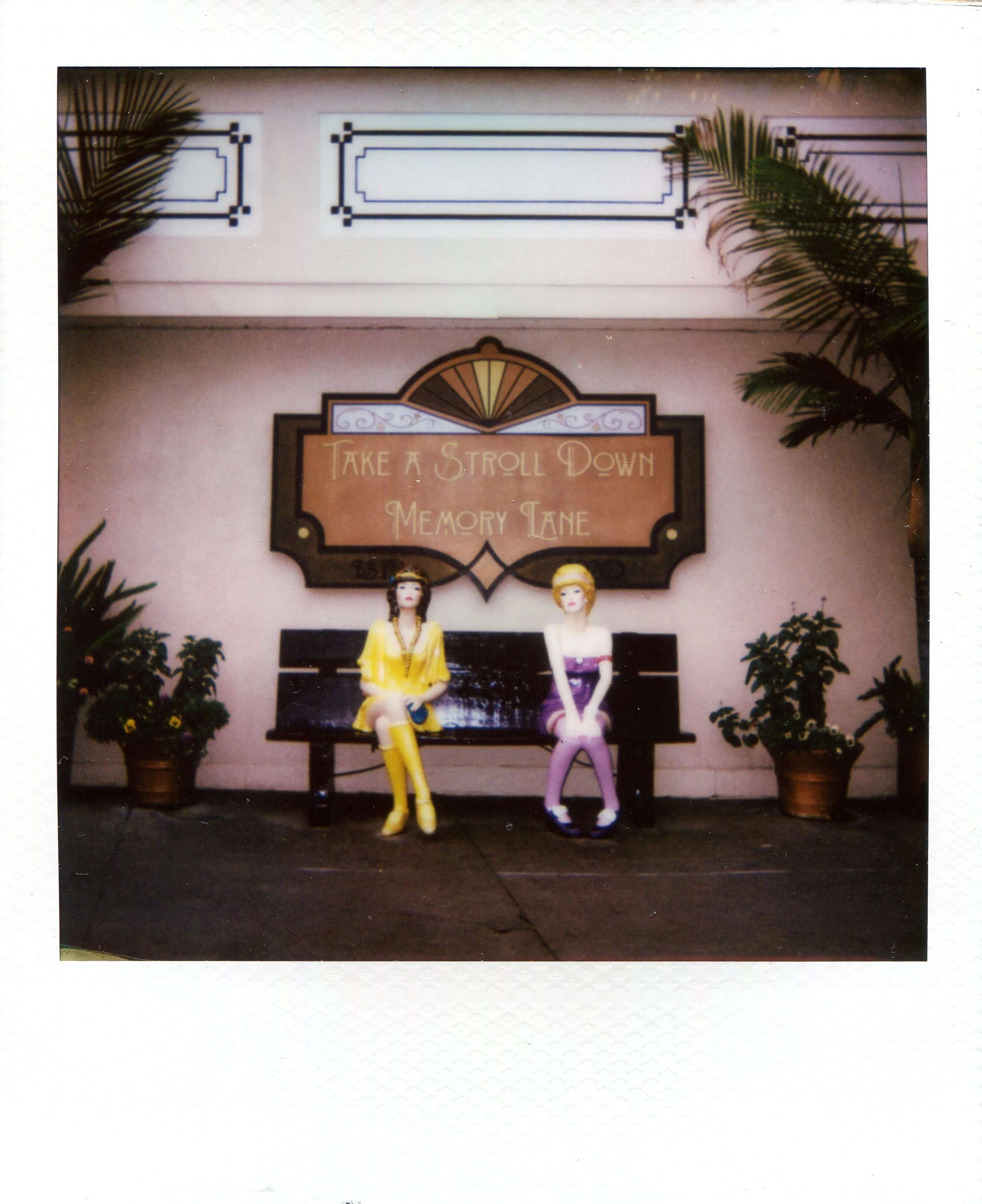 Two mannequins sitting on a park bench, dressed in colorful 1960s-style clothing, with a sign above them that says 'Take a Stroll Down Memory Lane,' flanked by potted plants on each side.