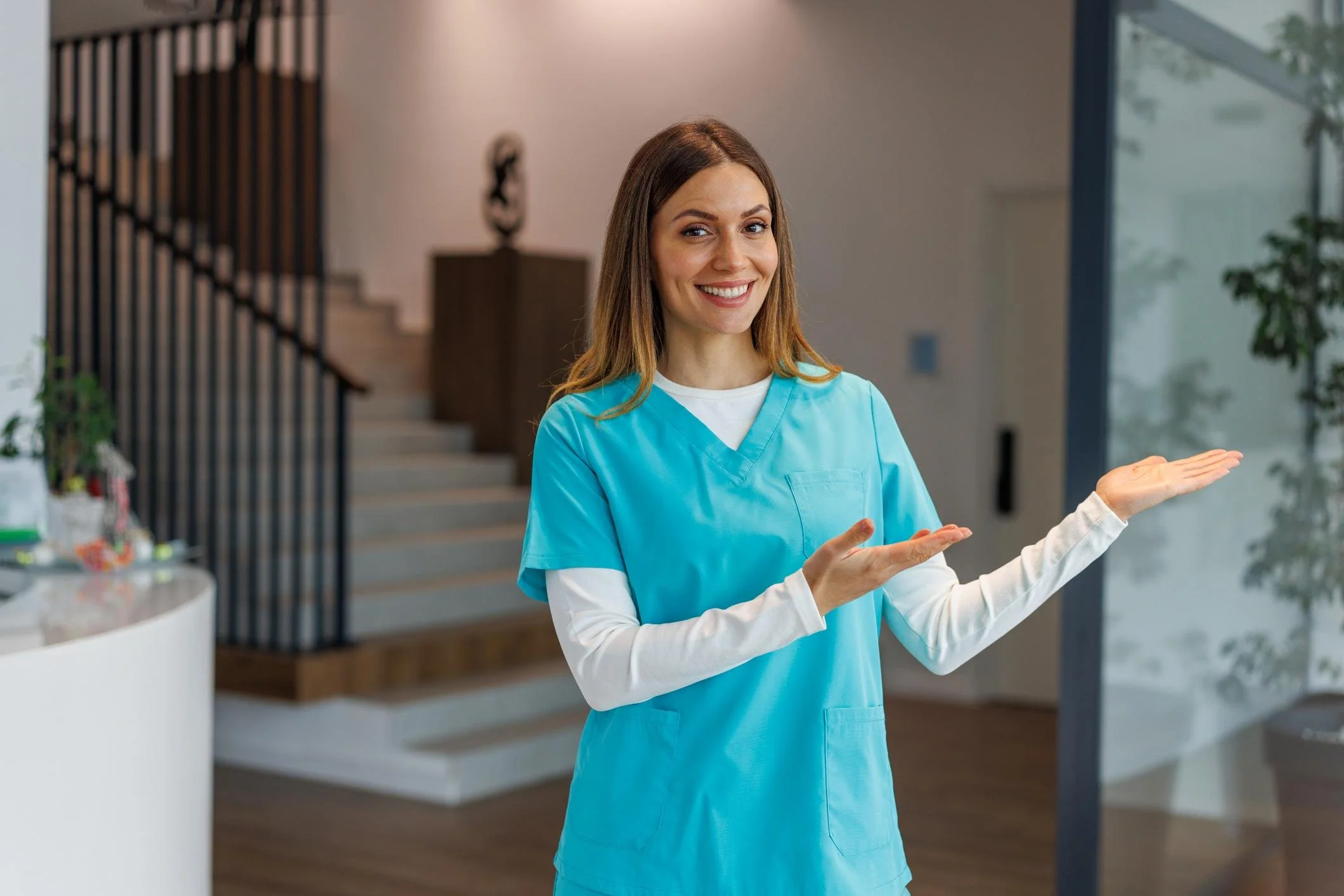 A smiling woman in blue scrubs standing in a modern indoor setting, gesturing with hands.