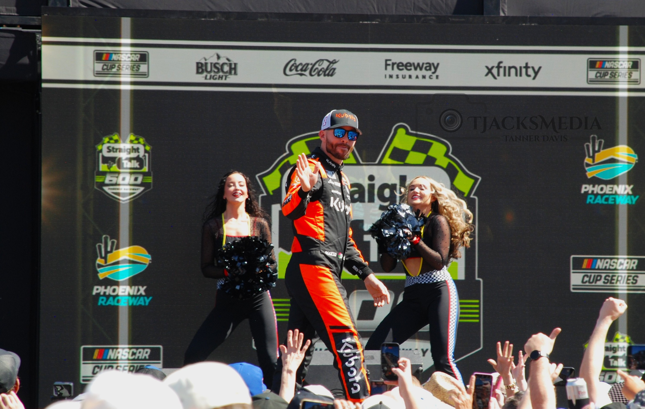 Race car driver on stage with two cheerleaders, waving and dancing, at Phoenix Raceway during a NASCAR event.