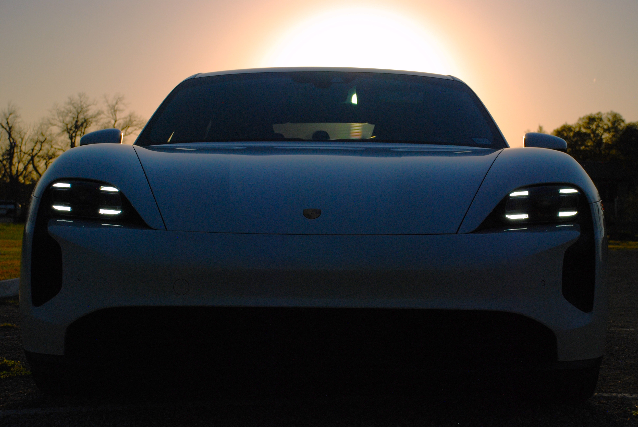 Front view of a white sports car with illuminated headlights, parked outdoors during sunset or sunrise with trees and sky in the background.