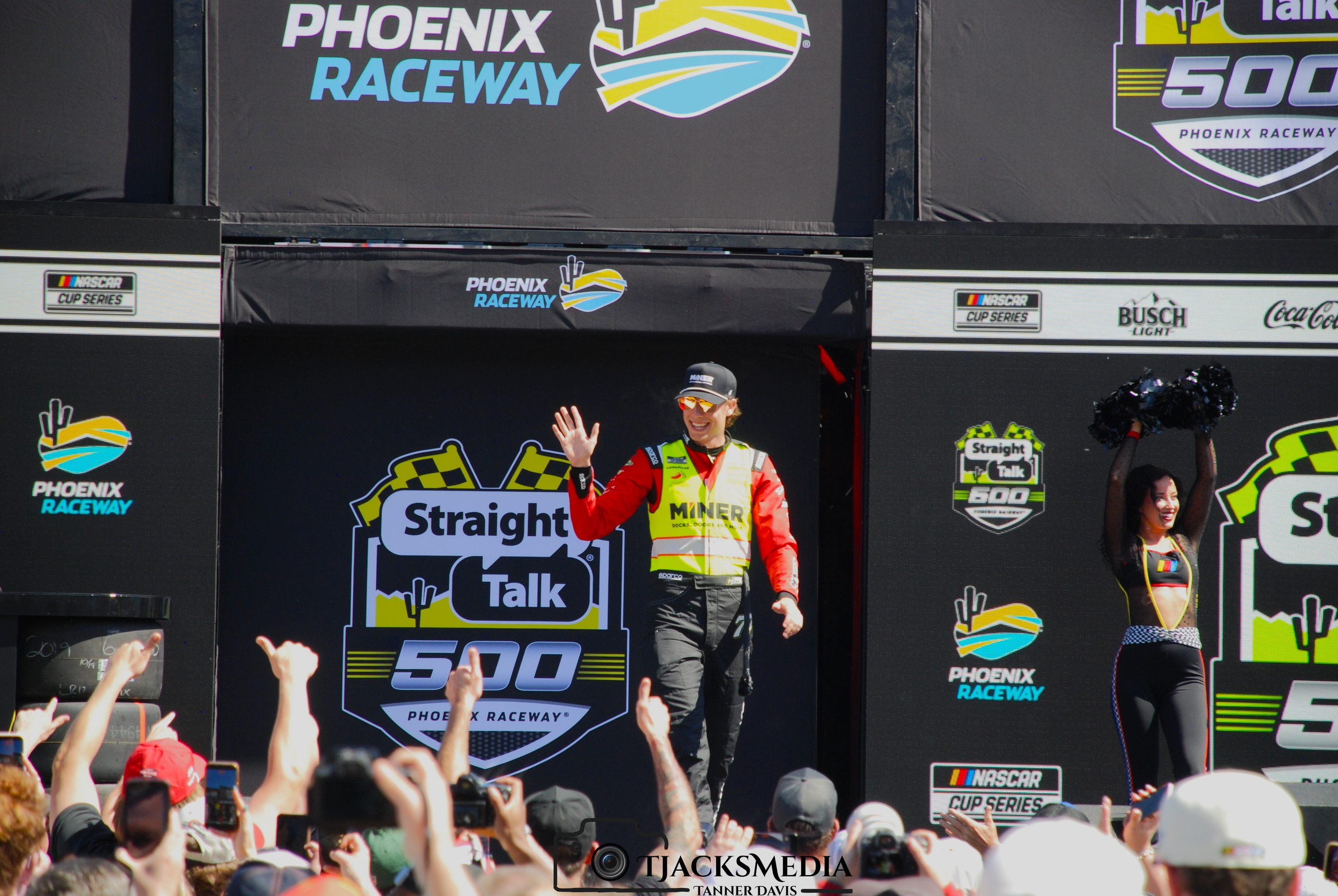 A race car driver on stage waving at the crowd at Phoenix Raceway during a Straight Talk 500 event, with a female cheerleader holding pom-poms beside him, in front of large event banners and logos.