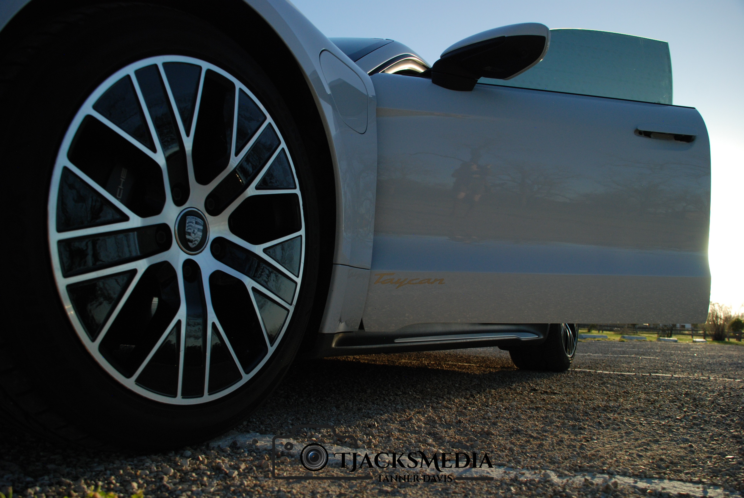 Close-up of the front wheel of a Porsche Taycan, parked on a gravel surface during sunset, with the car's side and a reflection of the sky visible.