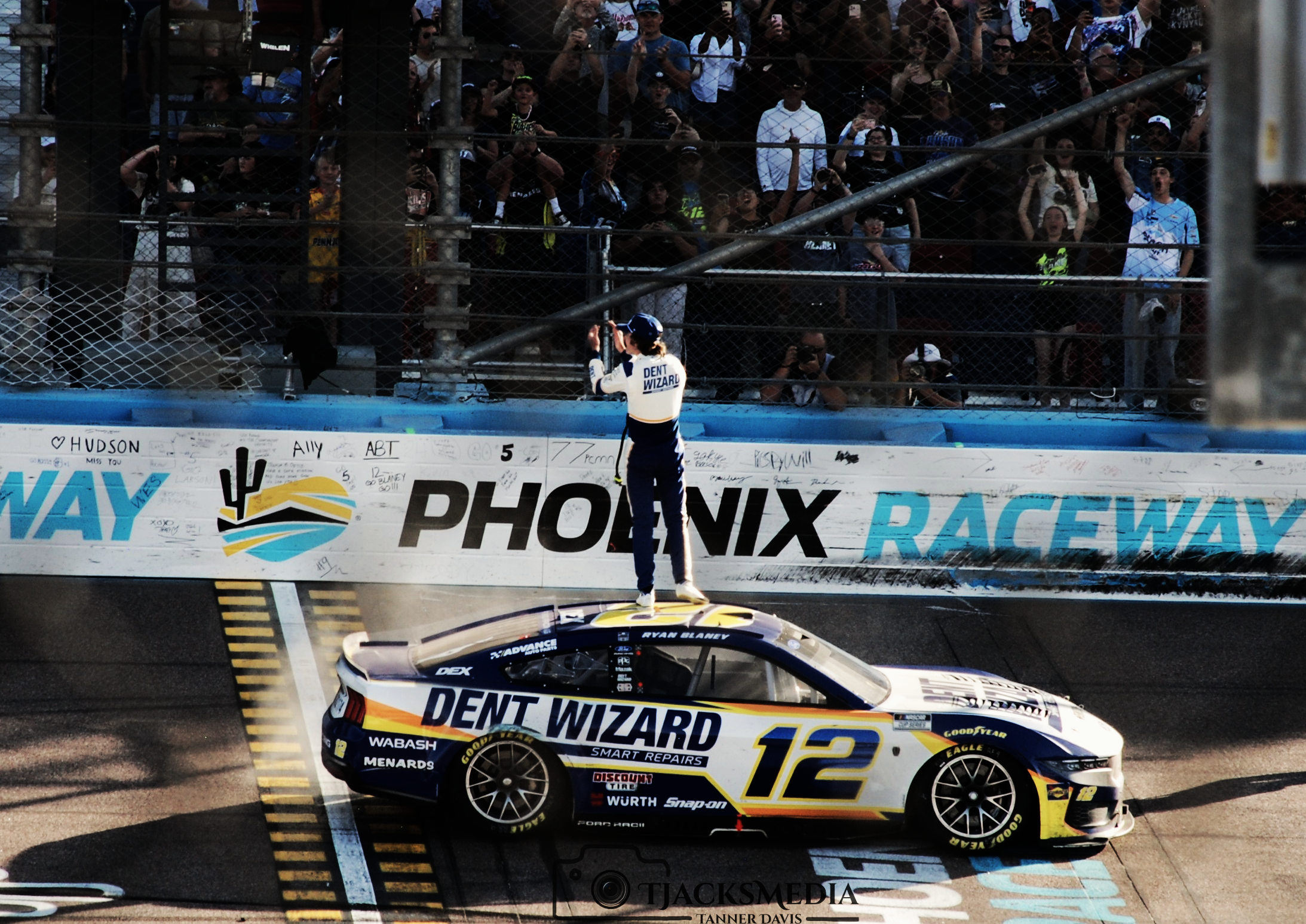 Race car with number 12 and sponsorship logos, person in racing gear standing on the roof, celebrating or gesturing, at Phoenix Raceway, crowd watching from stands behind a fence.
