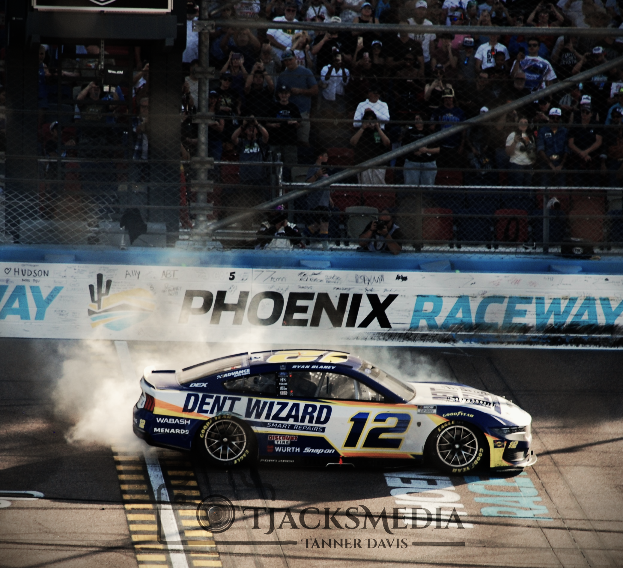 Race car number 12 with Dent Wizard sponsorship crosses the finish line at Phoenix Raceway, emitting smoke as fans watch from the stands.