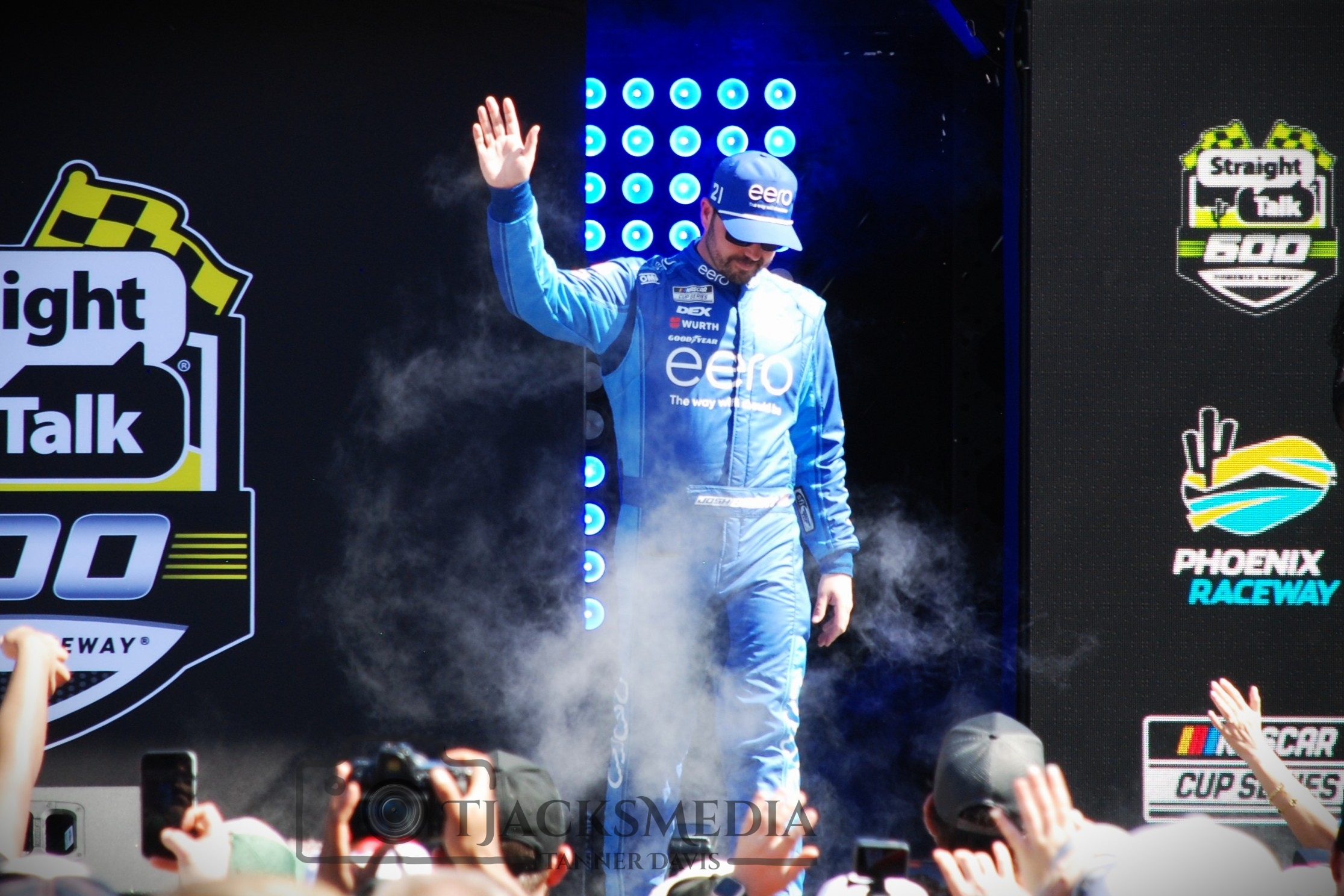 Race car driver in blue racing suit and cap on stage during a victory lap, waving to crowd at Phoenix Raceway, with smoke and cheering spectators taking photos.
