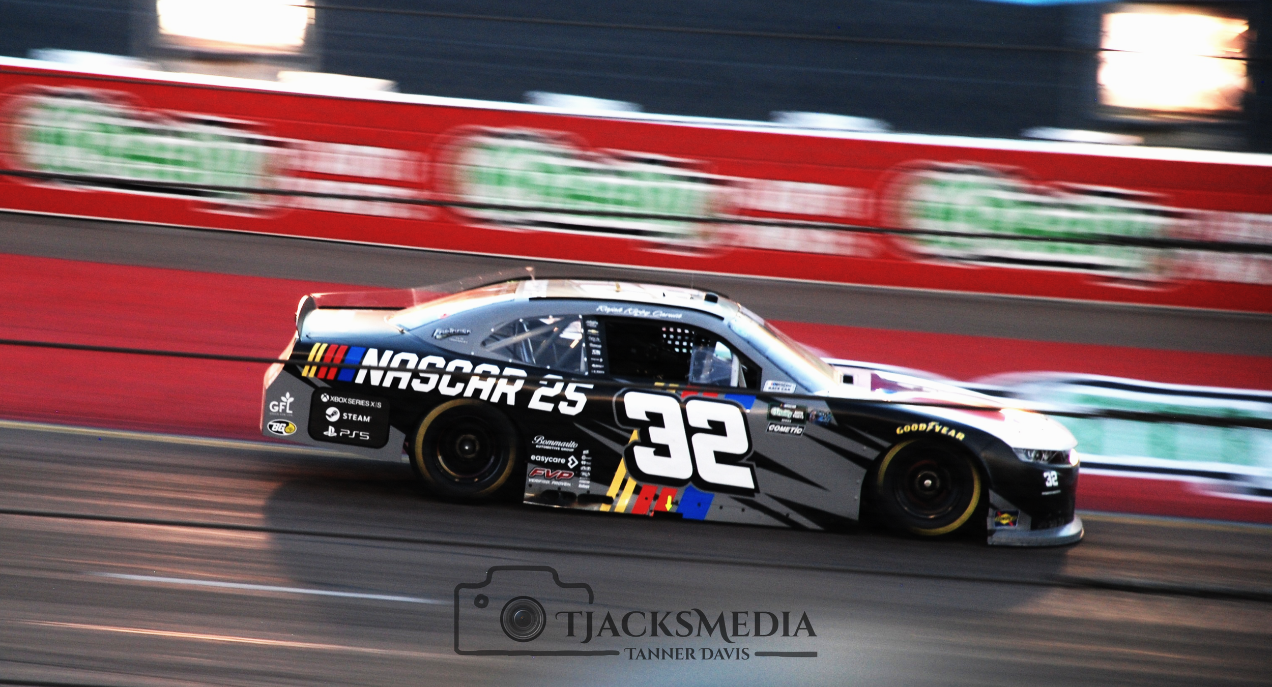 A black and gray NASCAR race car driven by Rajah Caruth with the number 32 speeding on a track, with a blurred red and black wall in the background.