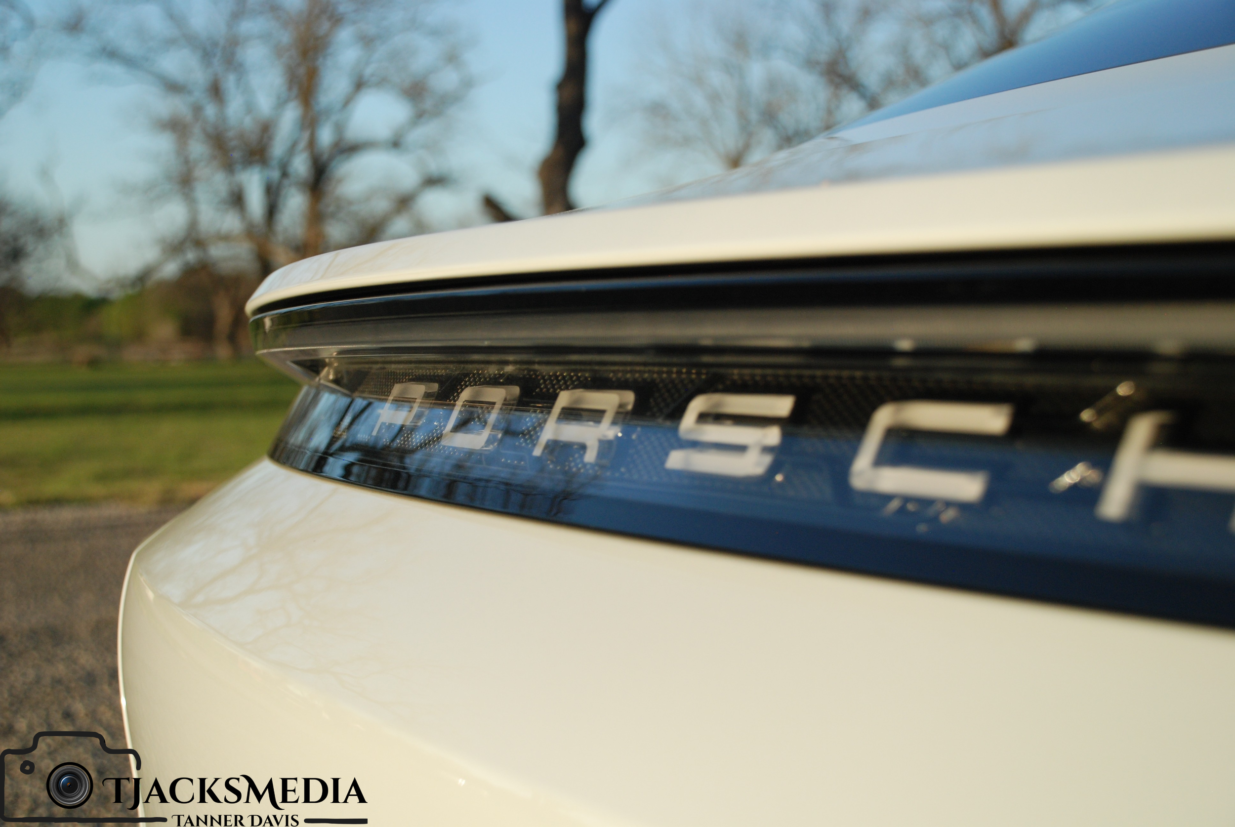 Close-up of the rear end of a white Porsche sports car, showing the Porsche emblem on the rear light, with trees in the background.