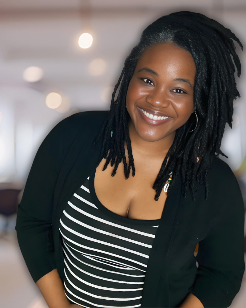 A woman with medium brown skin, black dreadlocks, and a bright smile, standing indoors with blurred lights in the background, wearing a black blazer over a black and white striped top.