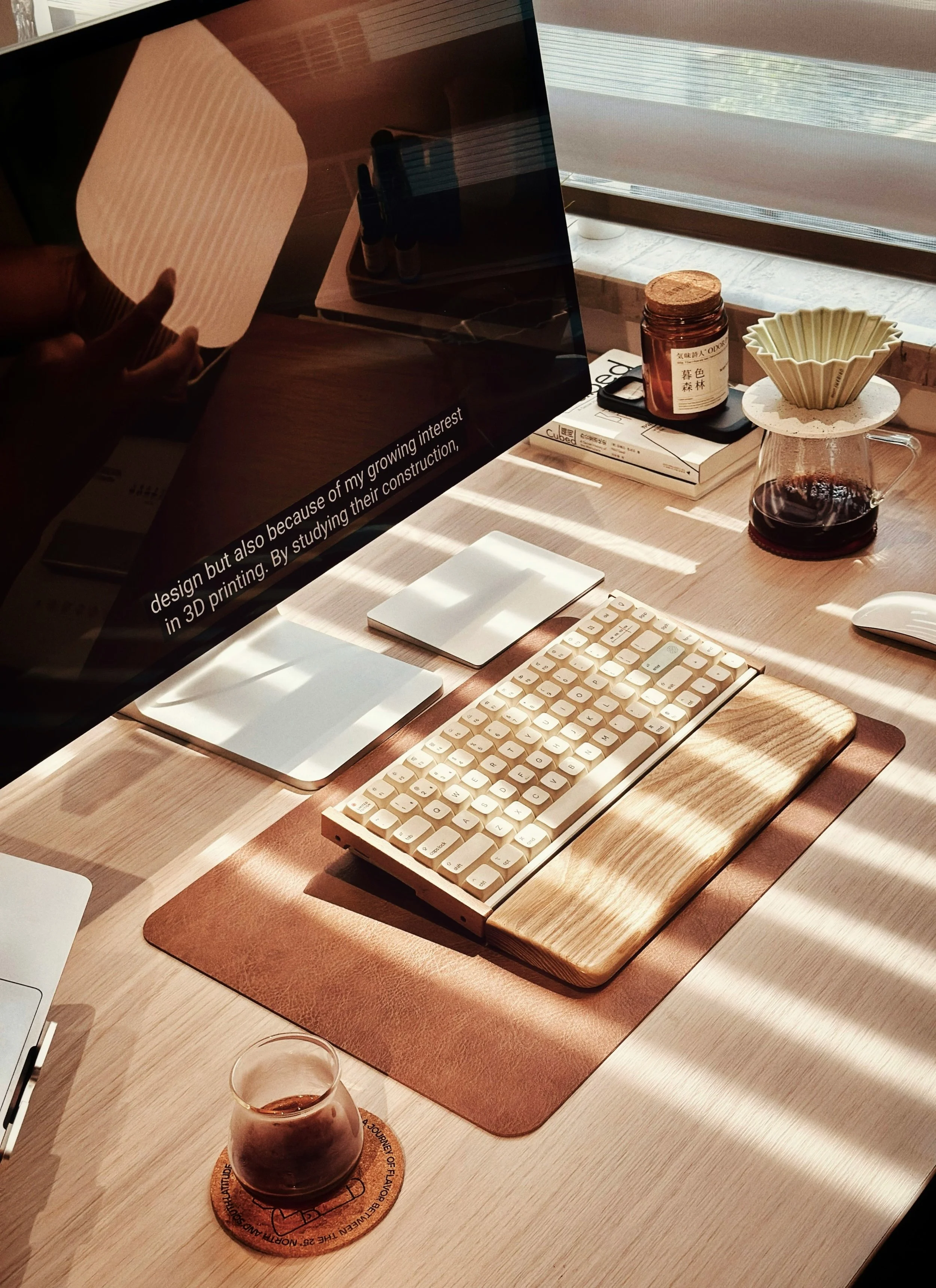 A workspace with a computer monitor, a white keyboard on a wooden desk mat, two white notepads, a glass of iced coffee, a coffee dripper with coffee underneath, a brown jar, a coffee mug, and some books near a window with sunlight streaming in.