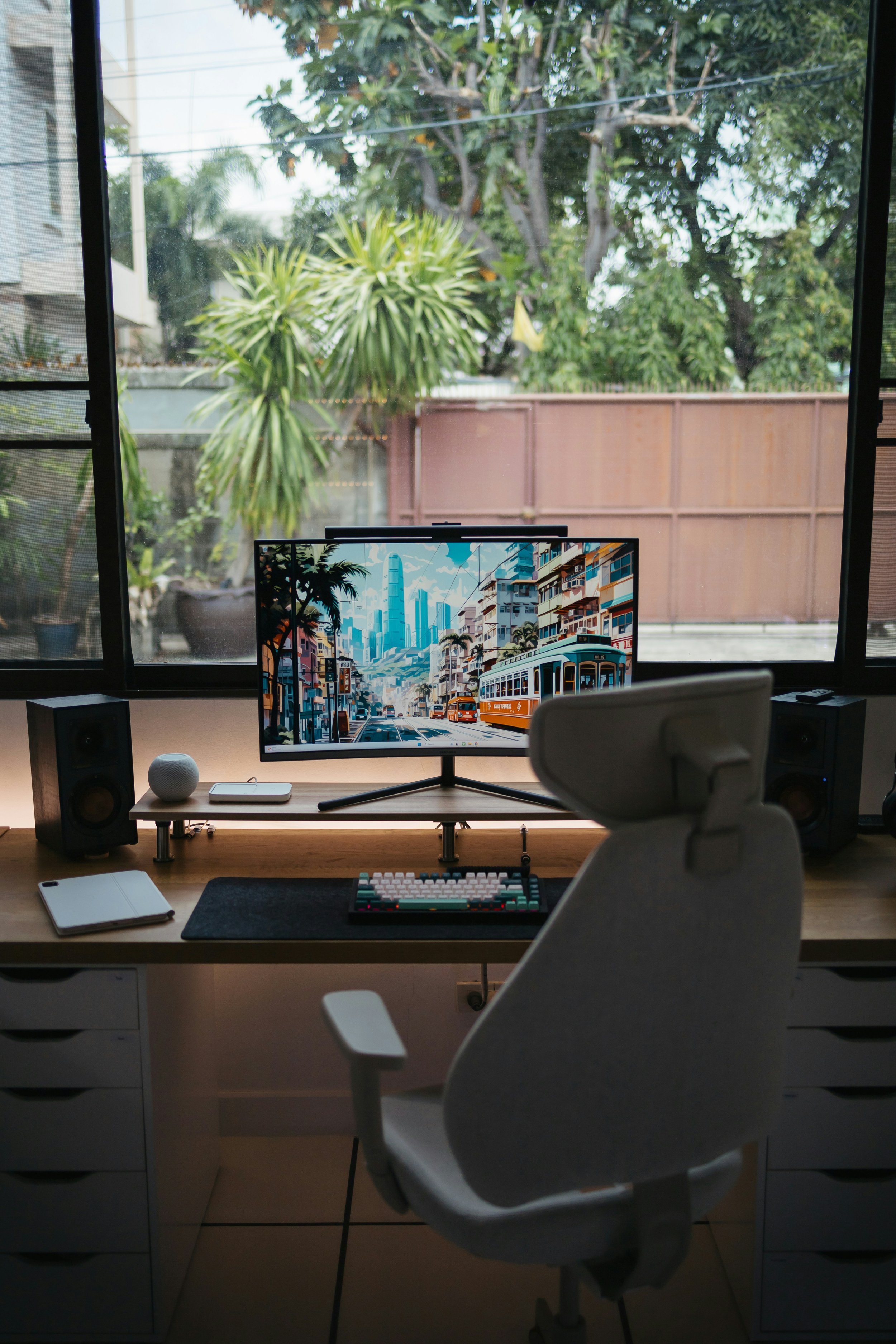 A modern home office setup with a large window showing greenery outside, a curved white ergonomic chair, a wooden desk with a computer monitor displaying a cityscape, a keyboard, a tablet, a speaker, and a small round object.