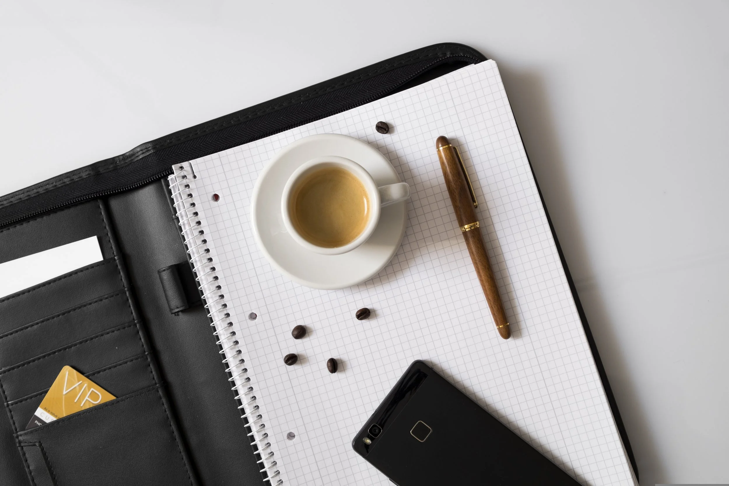 Open black leather zippered portfolio containing a white grid notebook, a small white cup of espresso on a saucer, a brown pen, a black smartphone, coffee beans, and a VIP card.