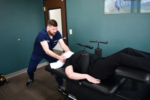 A chiropractor performs an adjustment on a patient lying face down on a treatment table in an office with teal walls.
